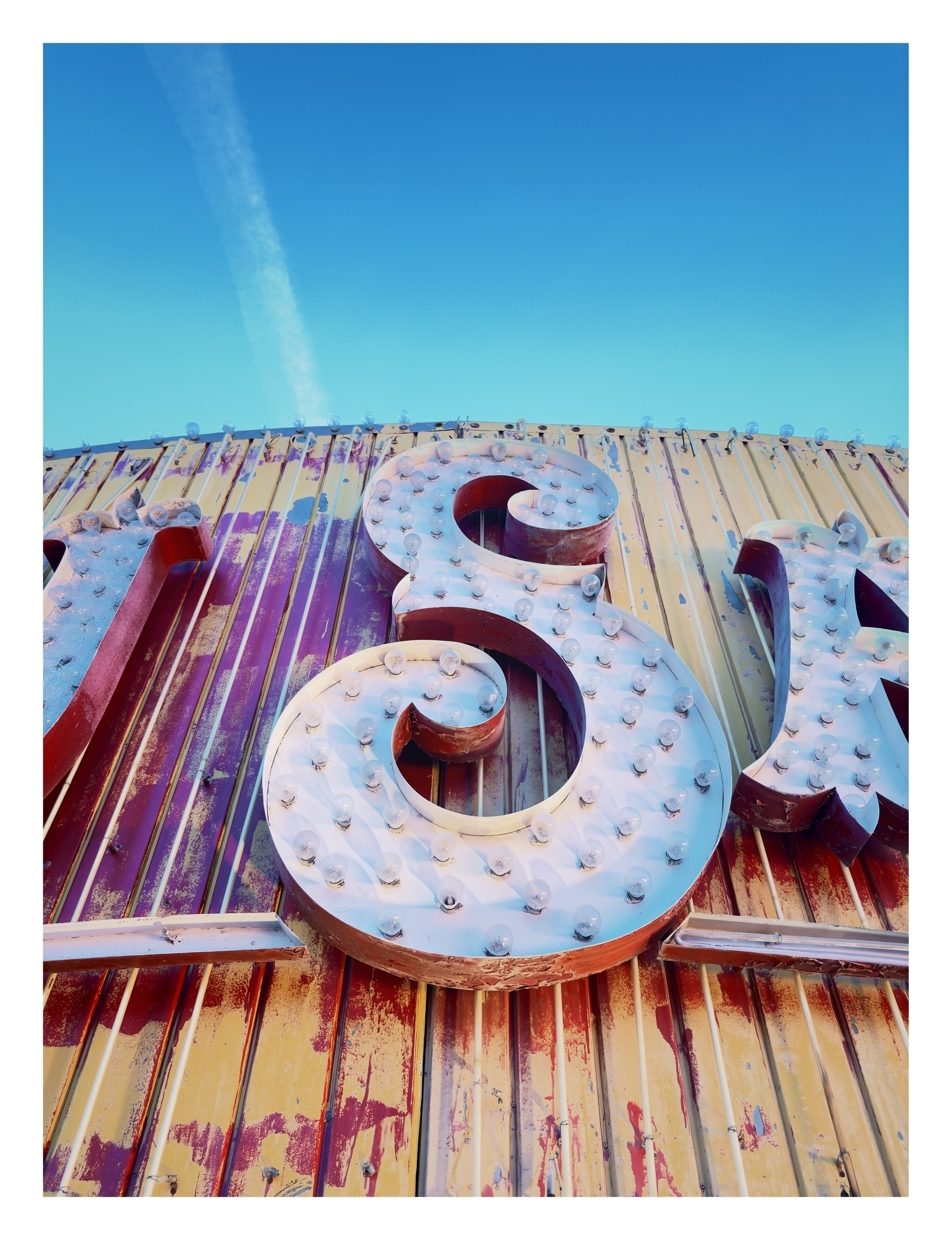 Large, illuminated letters affixed to a corrugated metal surface, under a clear blue sky with a contrail.