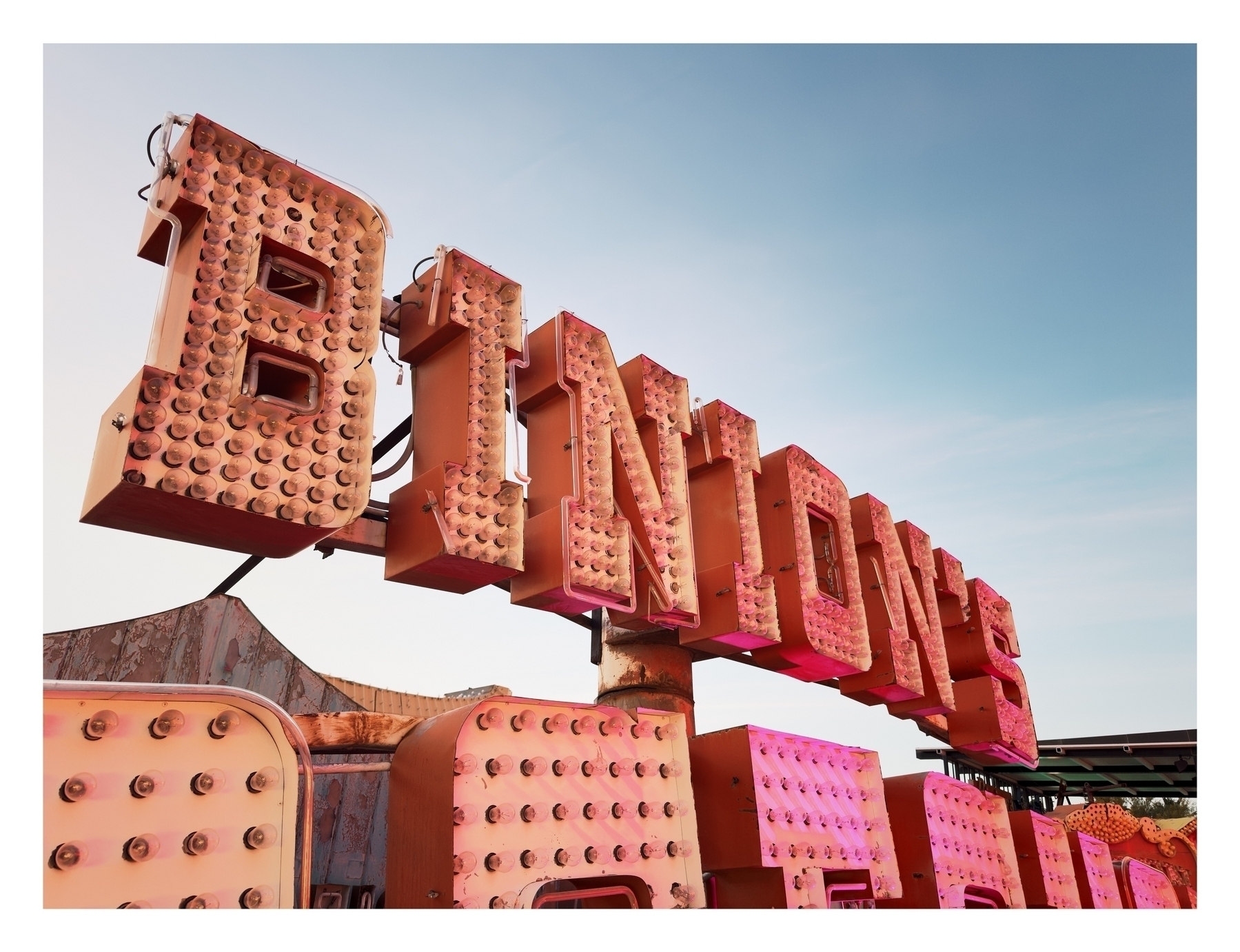 A weathered, pink neon sign spelling “BINION against a clear sky, mounted on rusted structures suggests a retro or abandoned setting.