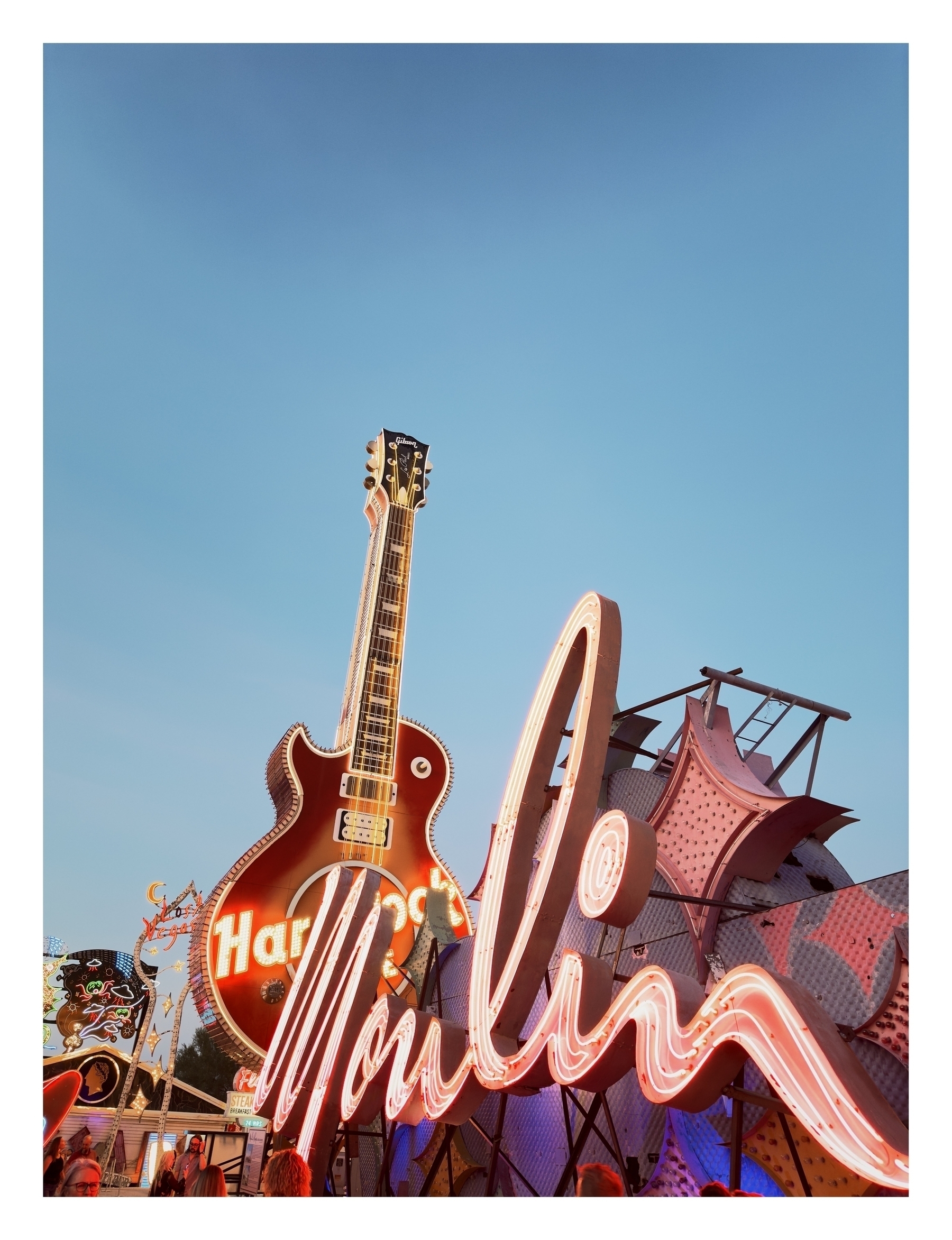 A large electric guitar sign with neon lights reads “Hard Rock” against a twilight sky, amidst other glowing amusement park attractions.