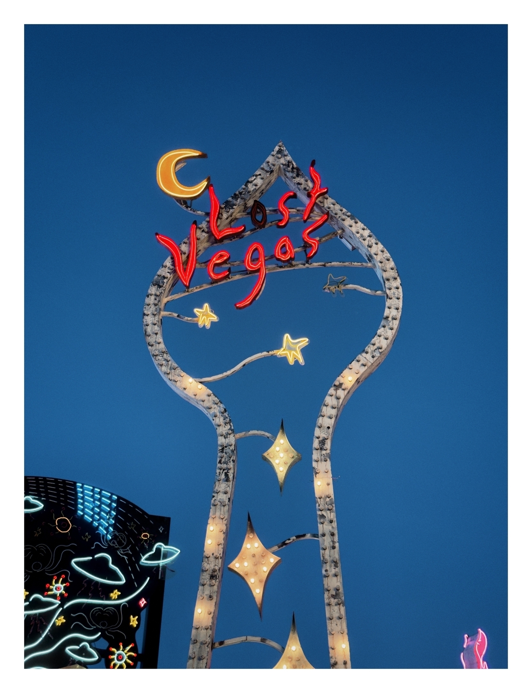 A neon sign reads “lost Vegas” atop a stylized structure, set against a blue sky. Below are illuminated stars and an animated building façade with neon graphics.