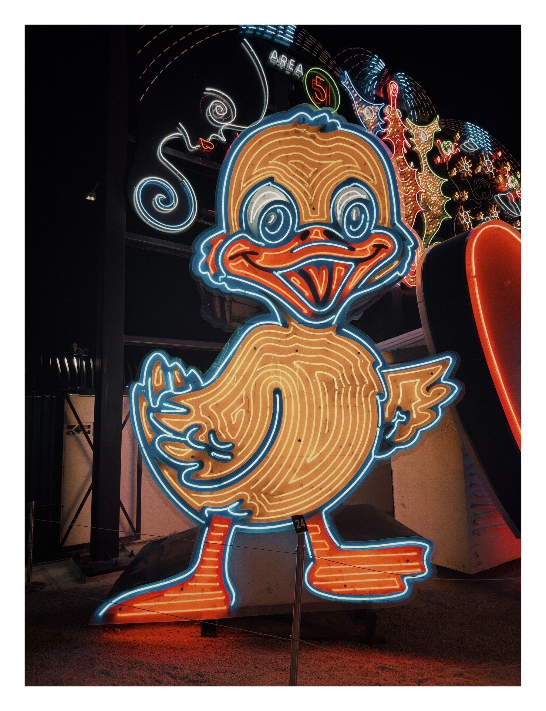 A neon-lit duck sign stands illuminated at night, providing a whimsical touch to a dark amusement park setting. No text visible.