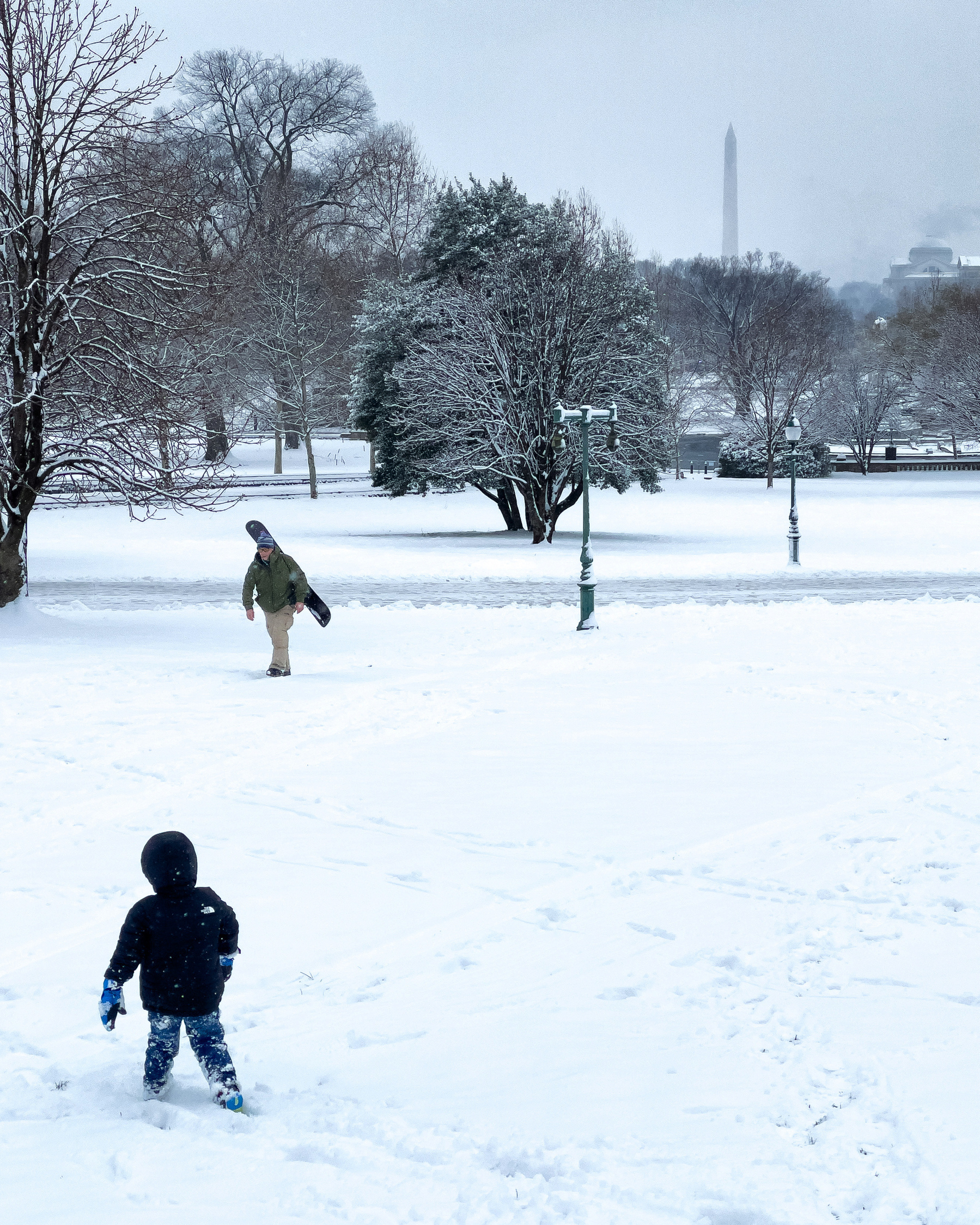 Taken from Capitol Hill, a small child’s back is to the viewer, a man with  a snowboard is walking towards us up a slight hill, and the Washington monument is visible in the distance. Everything is covered in snow.