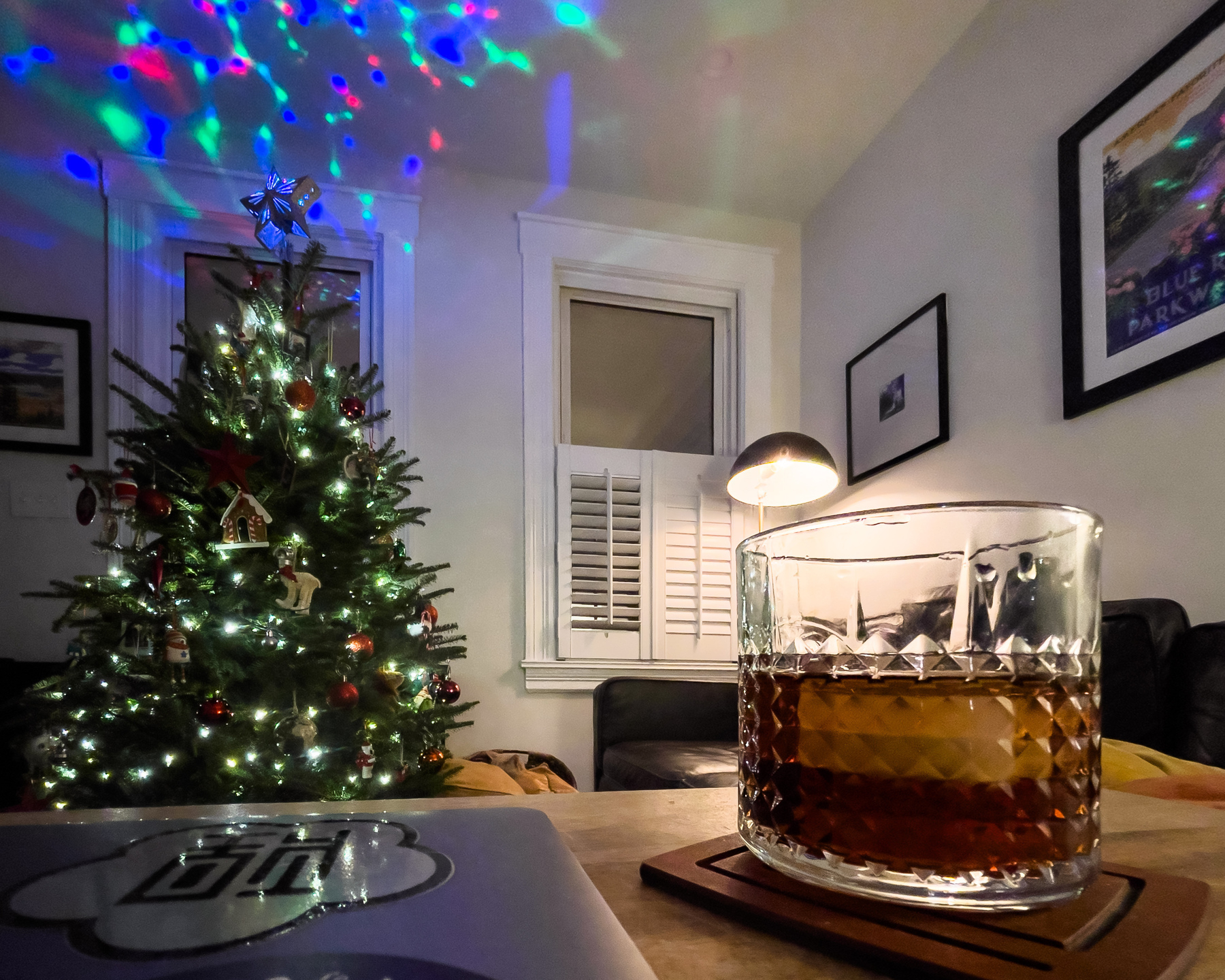 What appears to be a whisky cocktail in a rocks glass on a coaster, next to a closed sticker-laden laptop in the foreground. A Christmas tree projecting colorful lights into the ceiling and reflecting off of windows and framed art in the background.