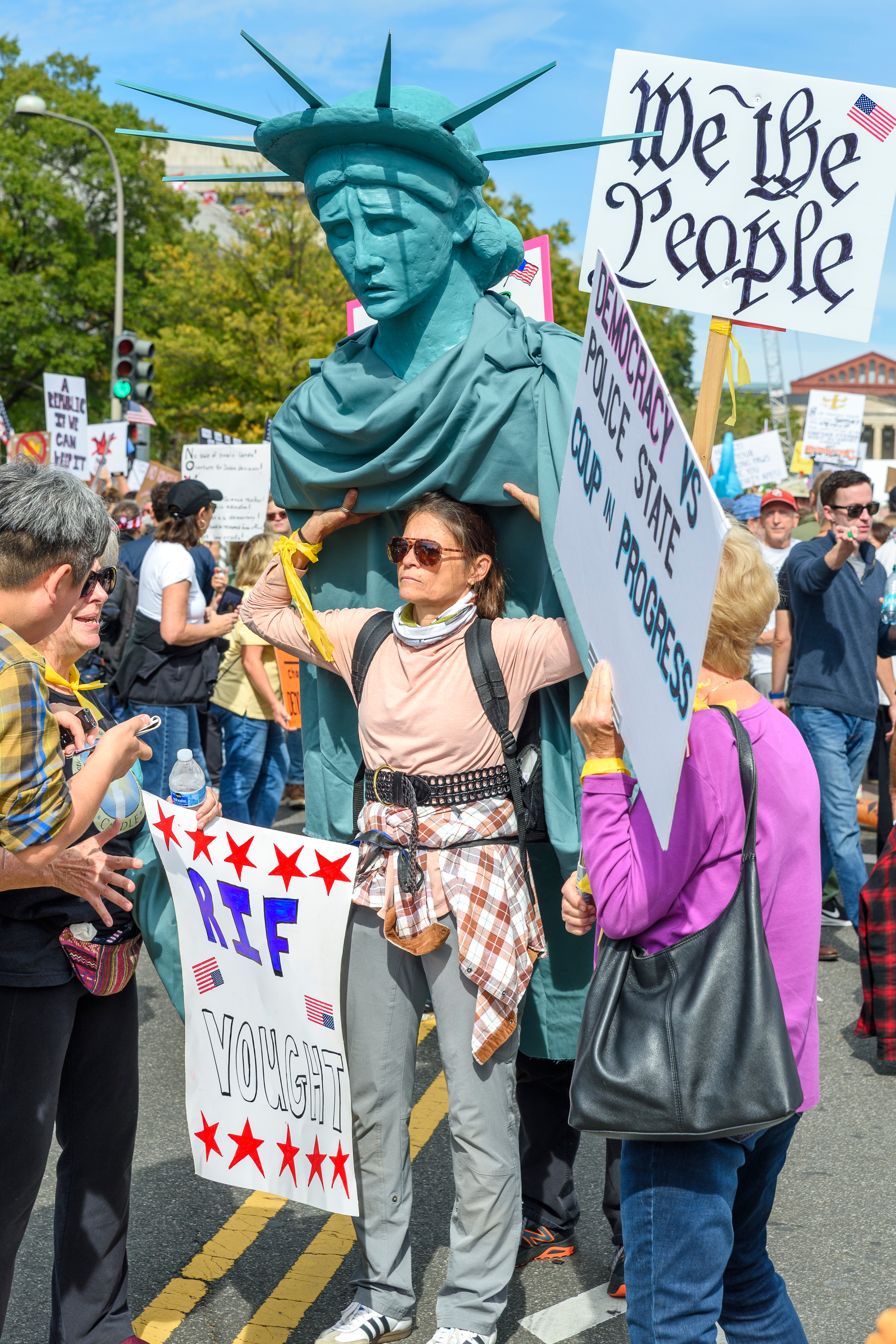 A woman holds a bust (possibly meant to be a worn worn) of the Statue of Liberty above herself. Lady Liberty has a look of dismay on her face. Next to her, a sign says We The People in Declaration-style script. A sign held by an onlooker says 'RIF Vought.'
