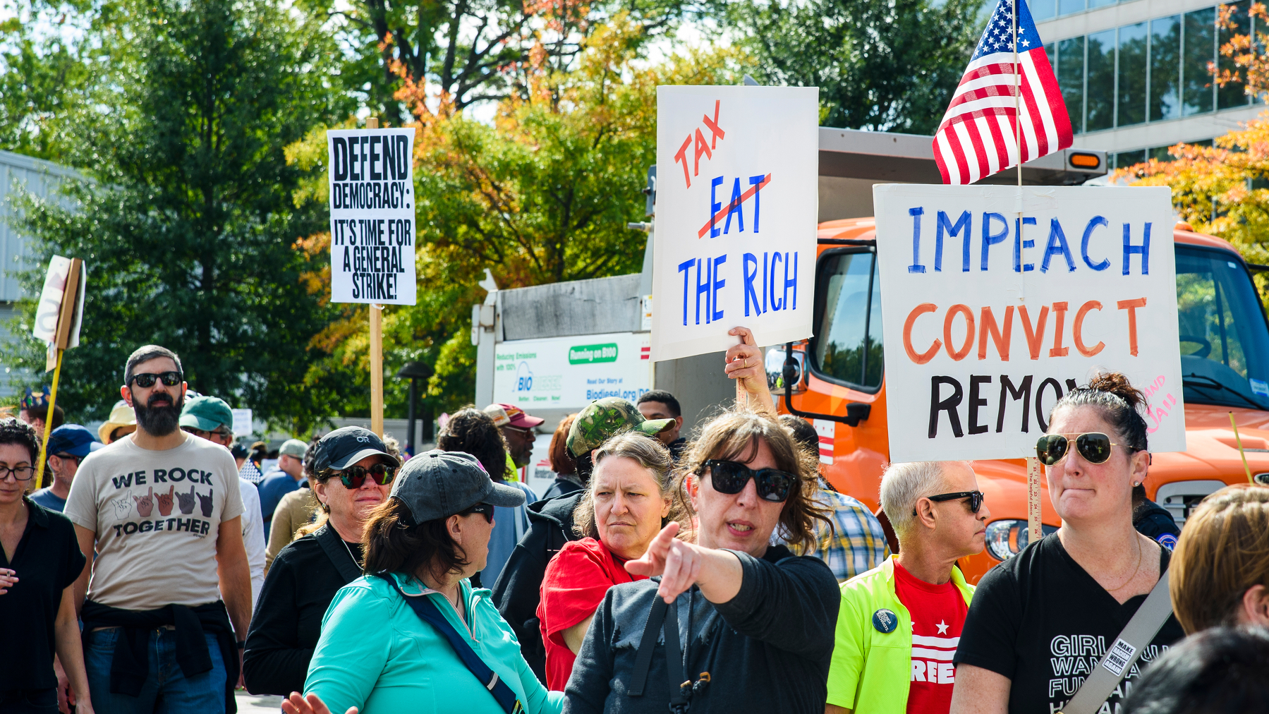 A wide shot of a folks at the No Kings rally today. Signs say 'Defend Democracy: It's Time For A General Strike', 'EAT (struck-through), TAX THE RICH', 'IMPEACH, CONVICT, REMOVE.' A free DC t-shirt can be seen, a man wears a shirt with multiple 'rock on' hand signs in different colors, it says 'we rock together.'