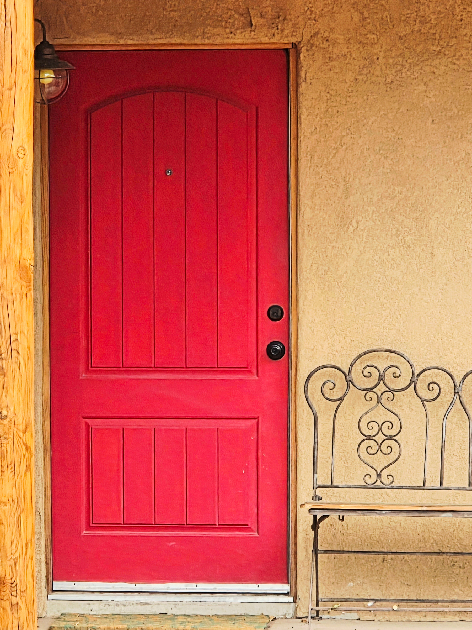 A red front door beside an old iron bench on an adobe porch