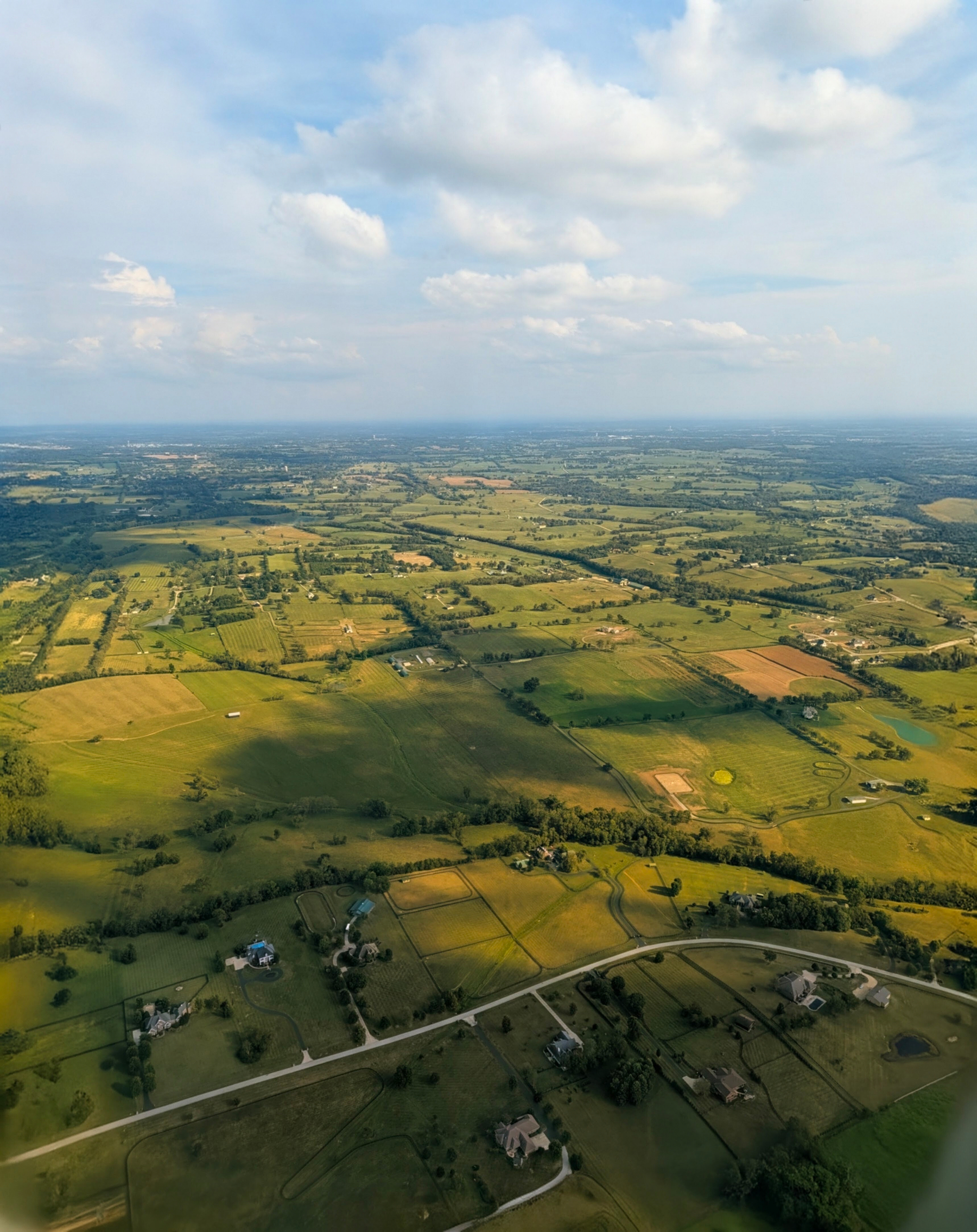 Auto-generated description: A vast rural landscape with fields, scattered houses, and roads stretches under a partly cloudy sky.