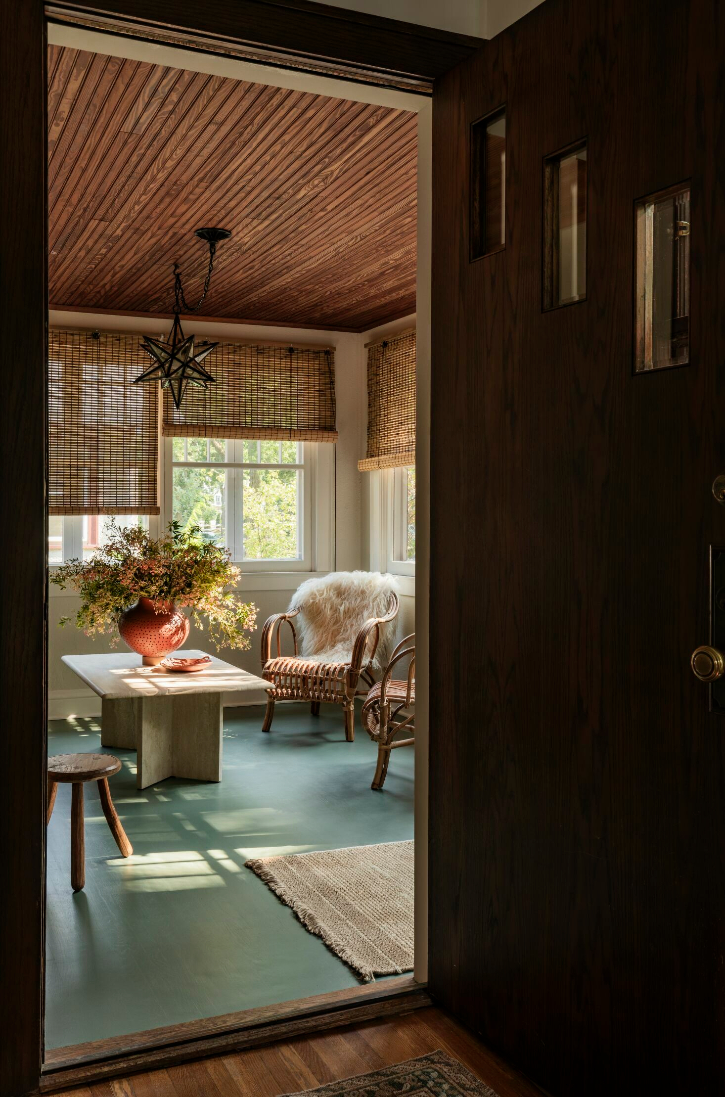 Cozy, rustic room features wooden ceilings, a wicker chair with a fur throw, a table with a large vase of flowers, and abundant natural light from windows with bamboo blinds.