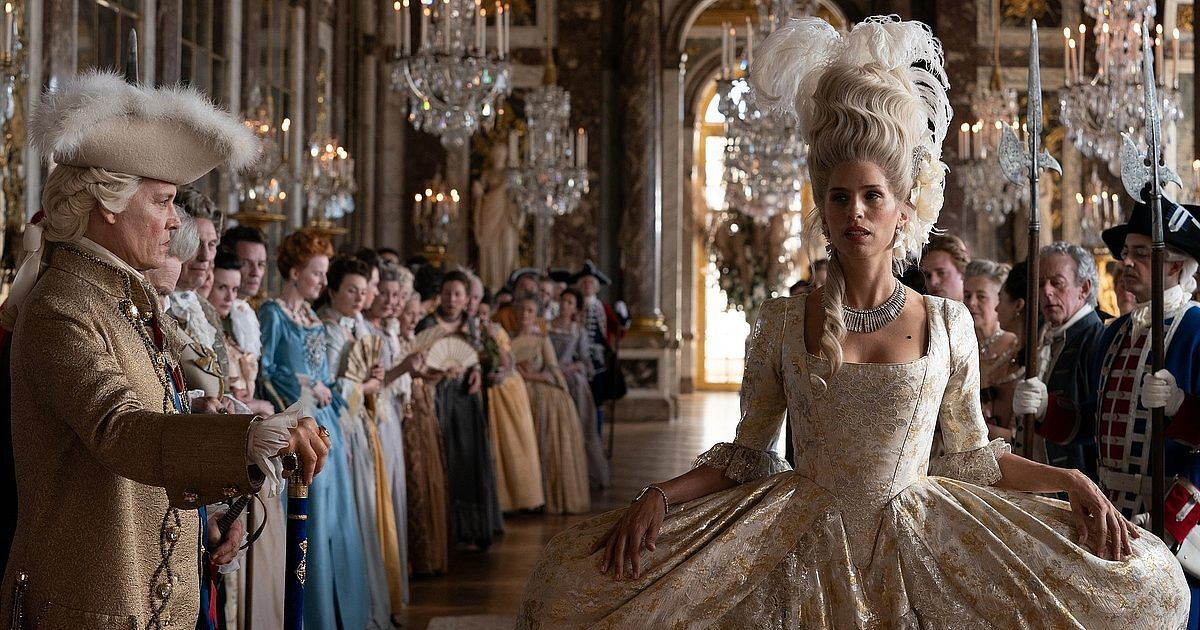 Auto-generated description: A woman in elaborate 18th-century attire stands in a luxurious, opulent room filled with people in period costumes.