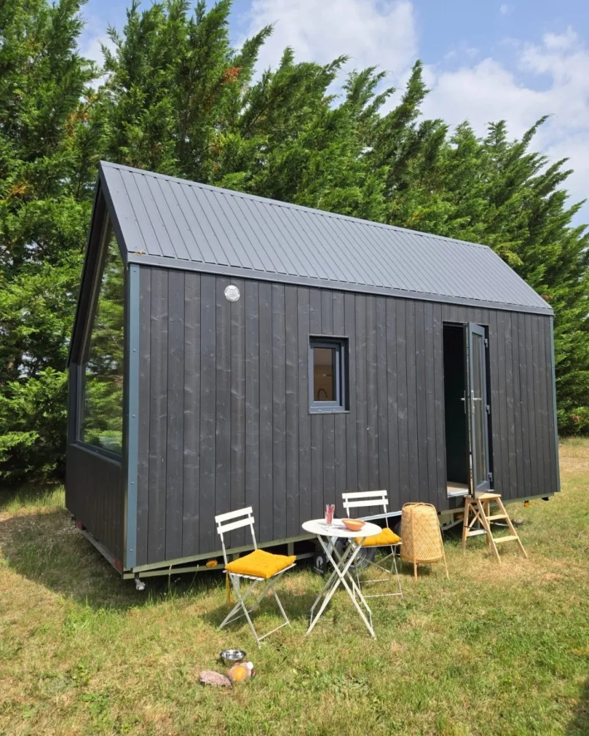 Auto-generated description: A modern tiny house with dark siding stands on a grassy area, accompanied by a small outdoor setup of a table and chairs.
