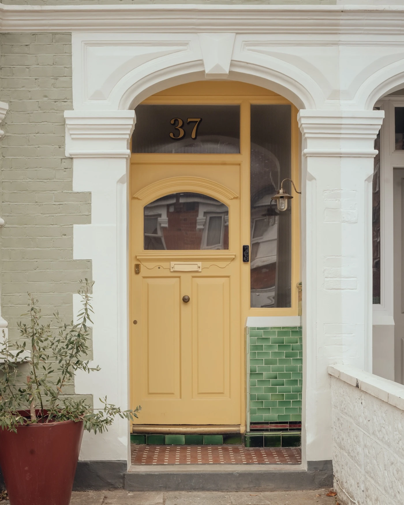 A yellow front door with the number 37 is set within a white archway, surrounded by green tiles and a potted plant