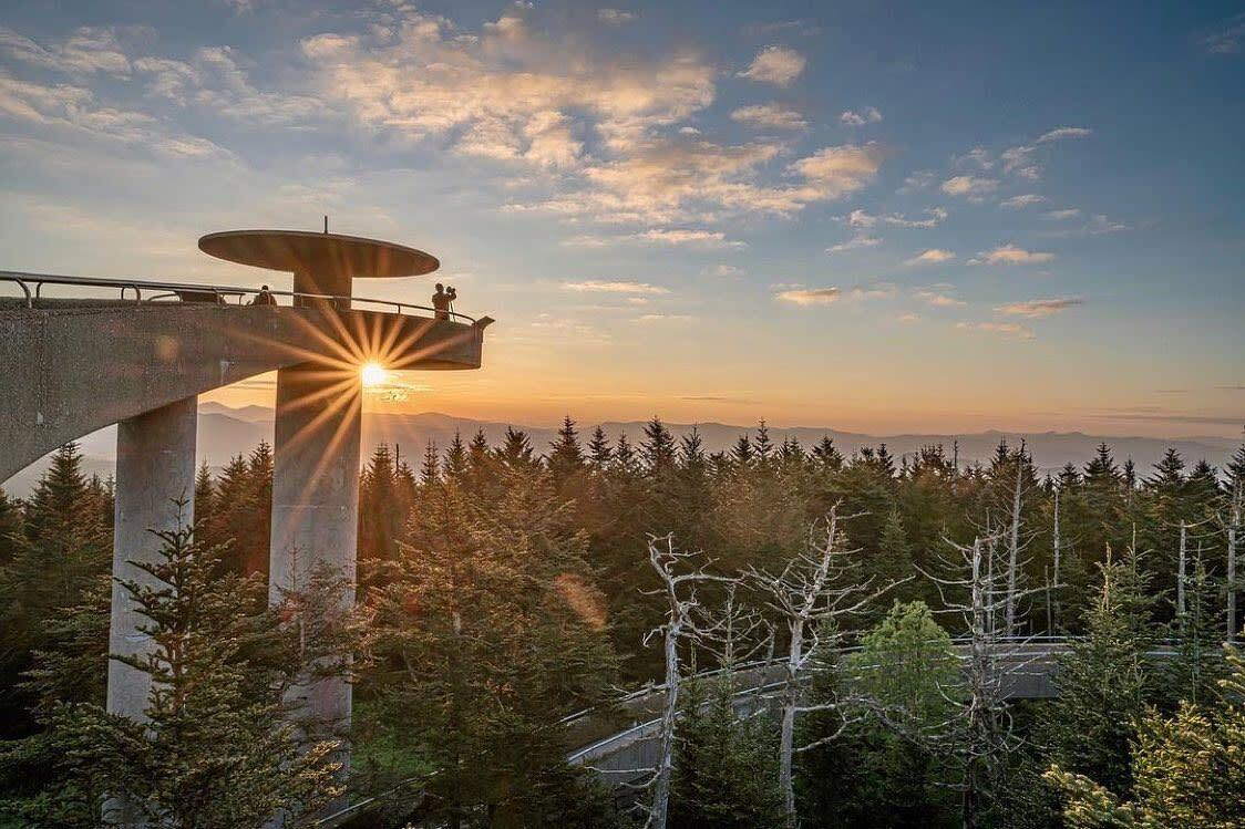 Auto-generated description: A scenic viewpoint structure rises above a forest with the sun setting behind it, casting a warm glow across the landscape.