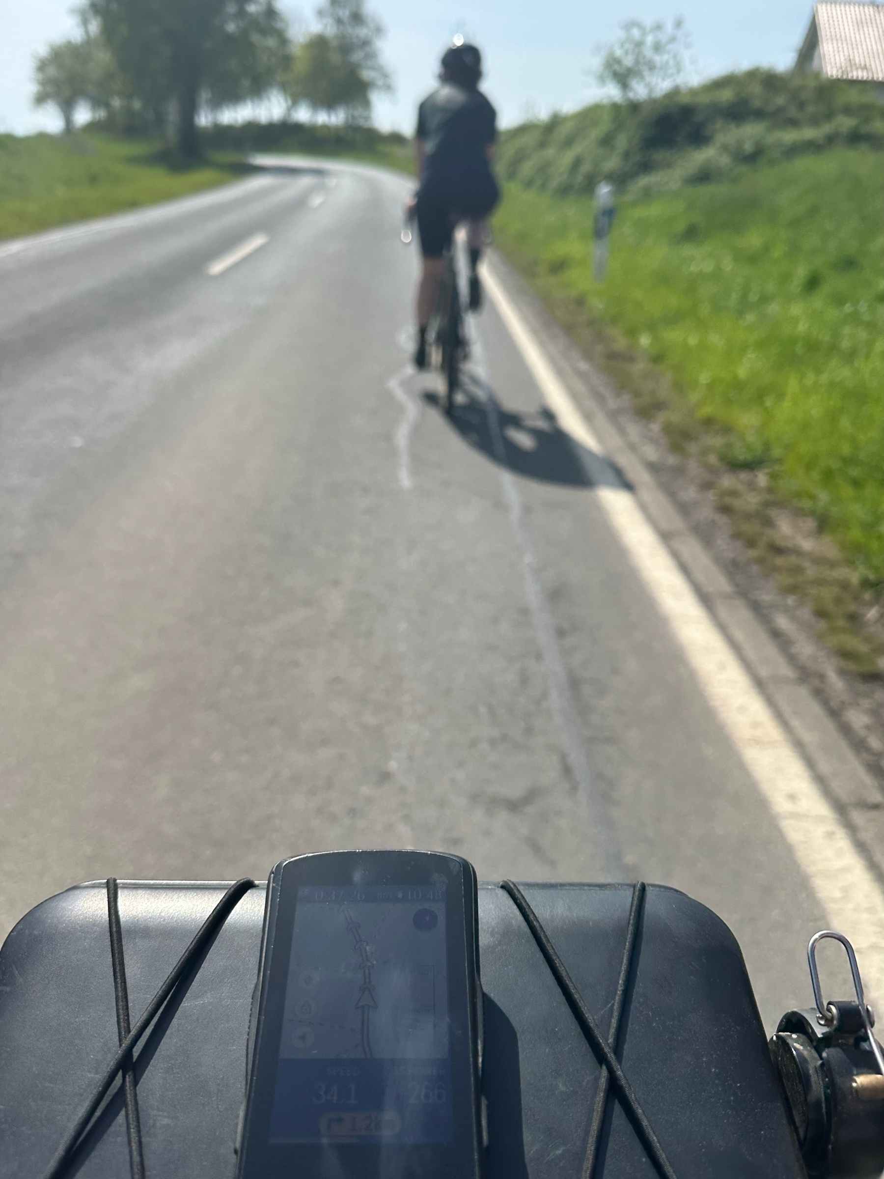 A cyclist is riding along a road, viewed from behind another cyclist's handlebars, displaying a cycling computer in clear weather.