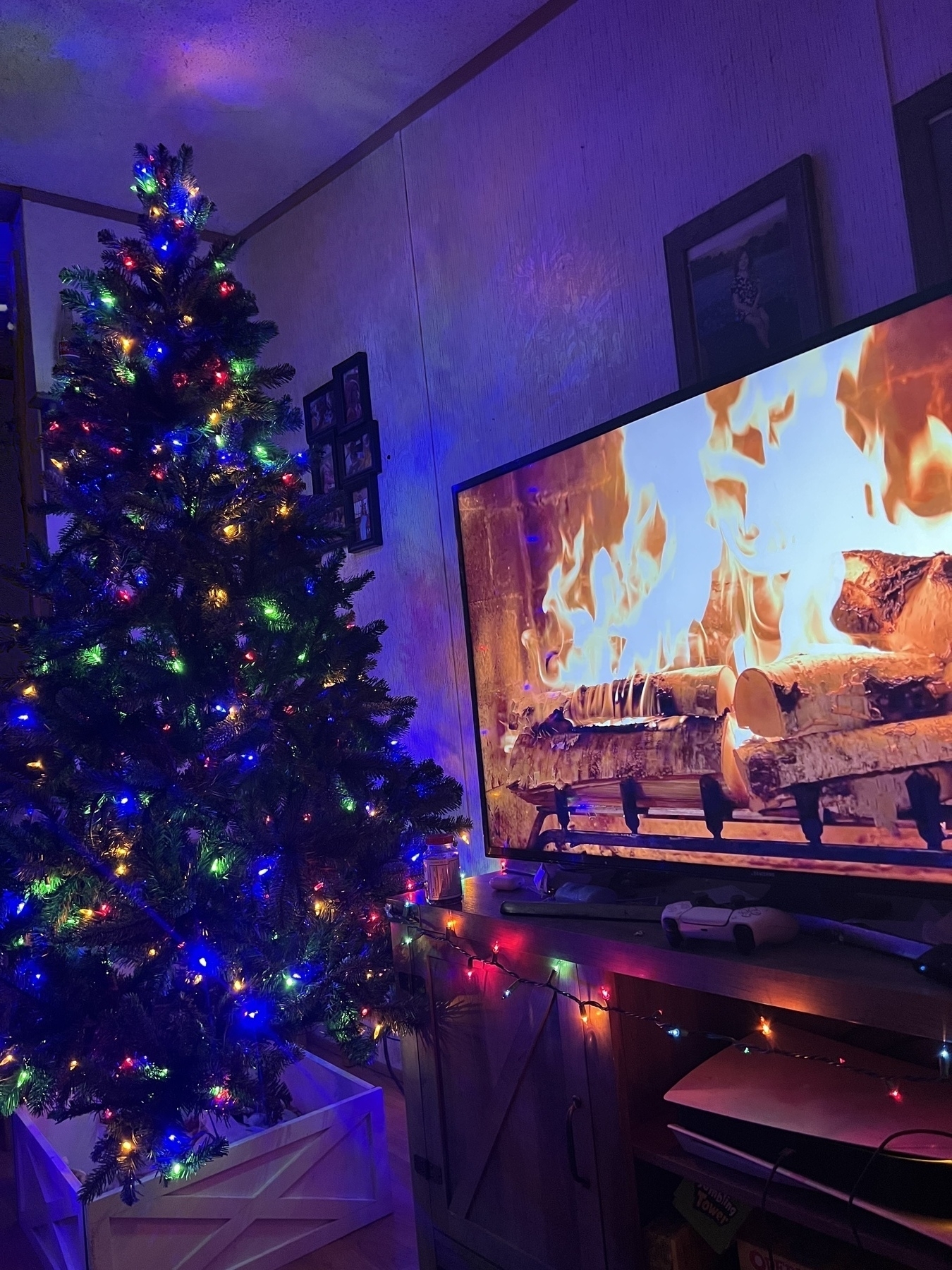 Christmas tree with multi color lights by a tv showing a fire place 