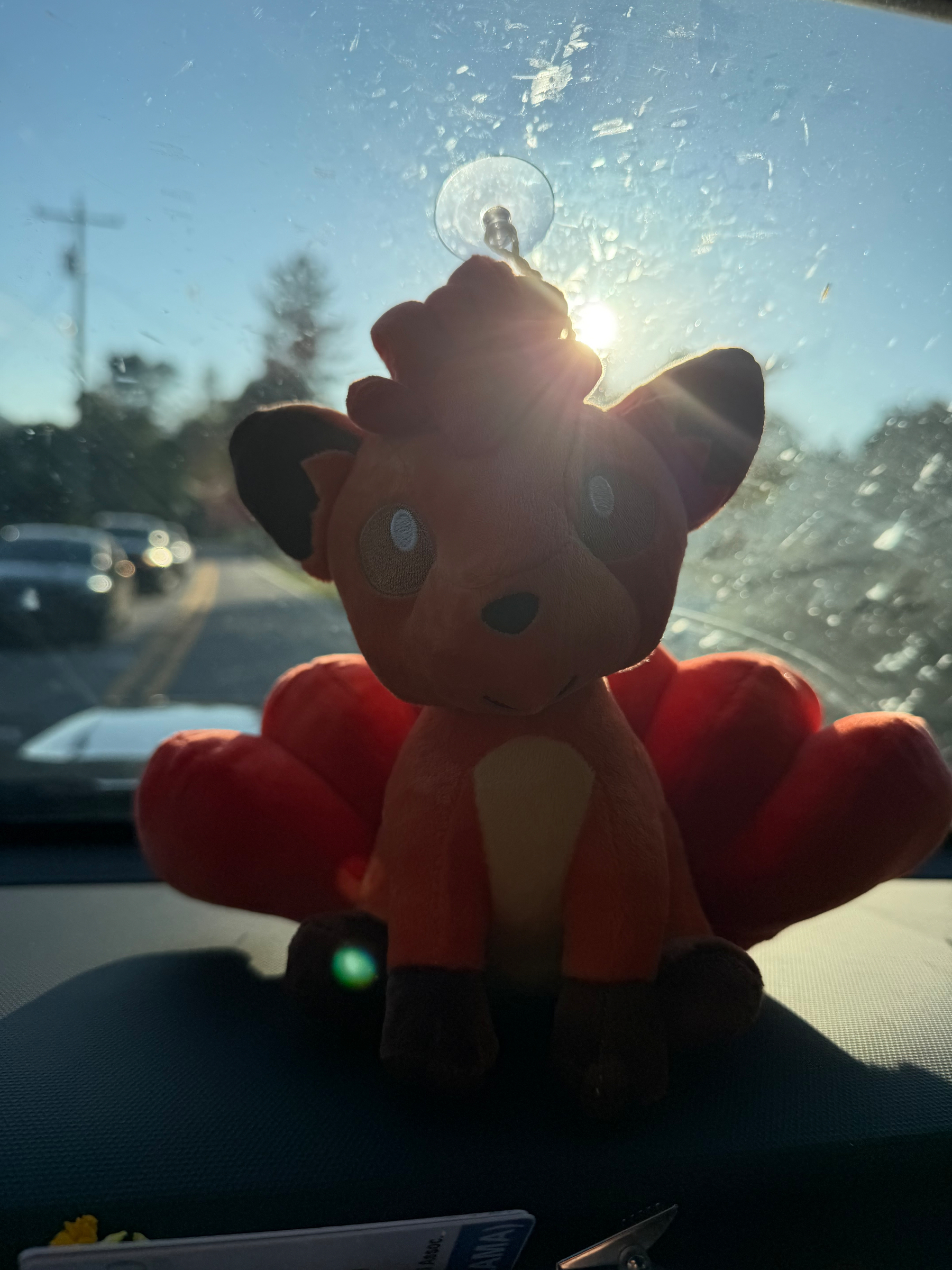 A plush toy with fox-like features is placed on a car dashboard with sunlight streaming through the windshield.