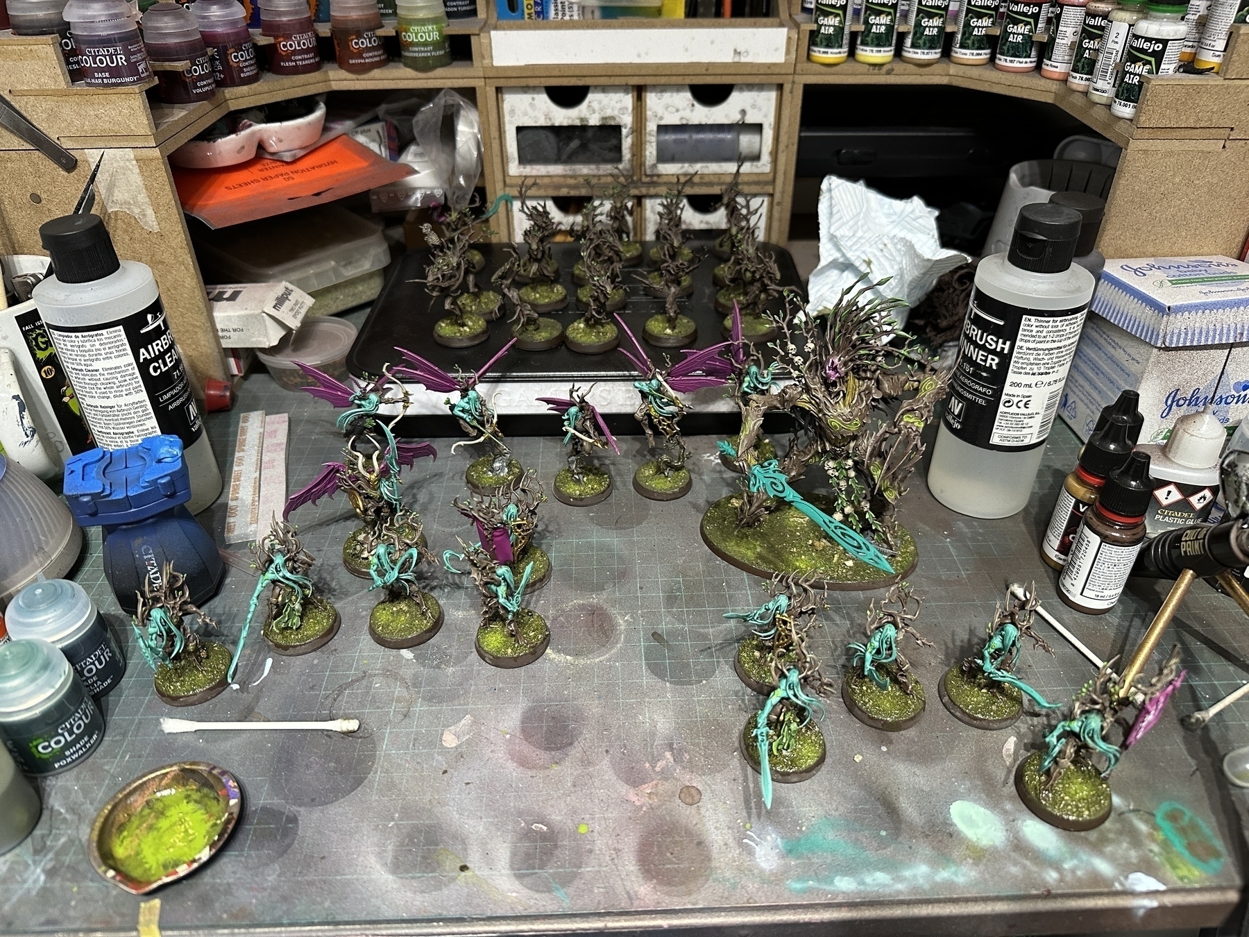 A collection of mostly painted miniatures from the Sylvaneth faction of Age of Sigmar, on a messy desk. The primary colours are woody browns, cold jade greens, warm forest greens, plus some magenta/pinks and gold as “spot” colours
