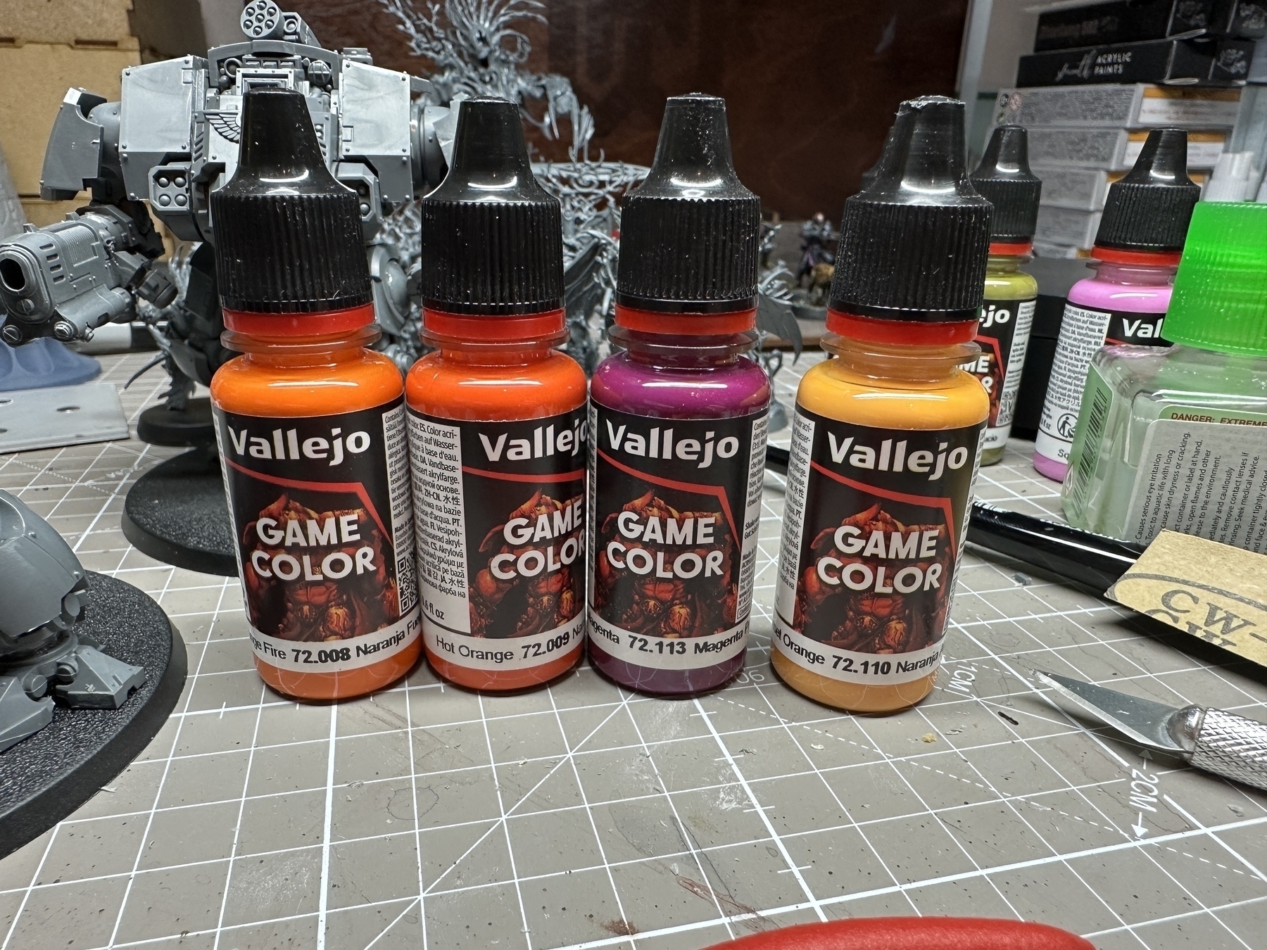 4 Vallejo Game Color paint bottles. Left to right they are: Orange Fire, a bold orange mid-tone; Hot Orange, a bold orange that leans to red; Deep Magenta, which is a darker, slightly purple magenta tone; Sunset Orange, a lighter orange that leans more to yellow