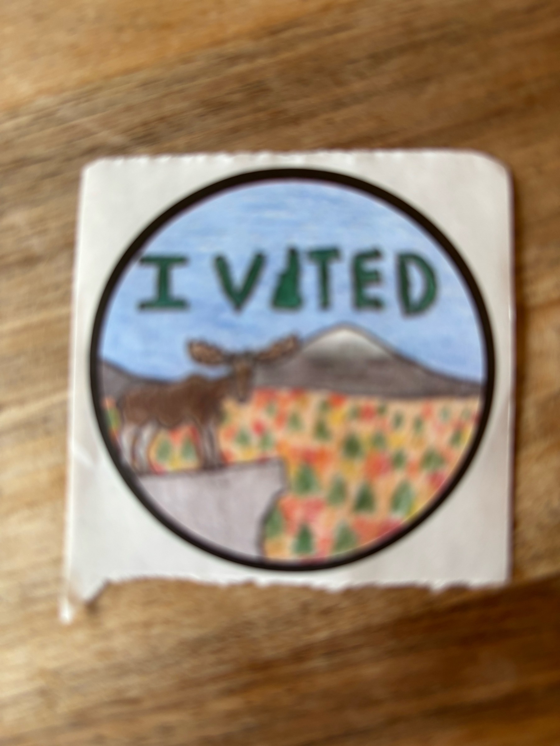 A fuzzy picture of a round sticker with a hand-drawn childlike picture of a moose, a blue sky, and the words "I Voted"