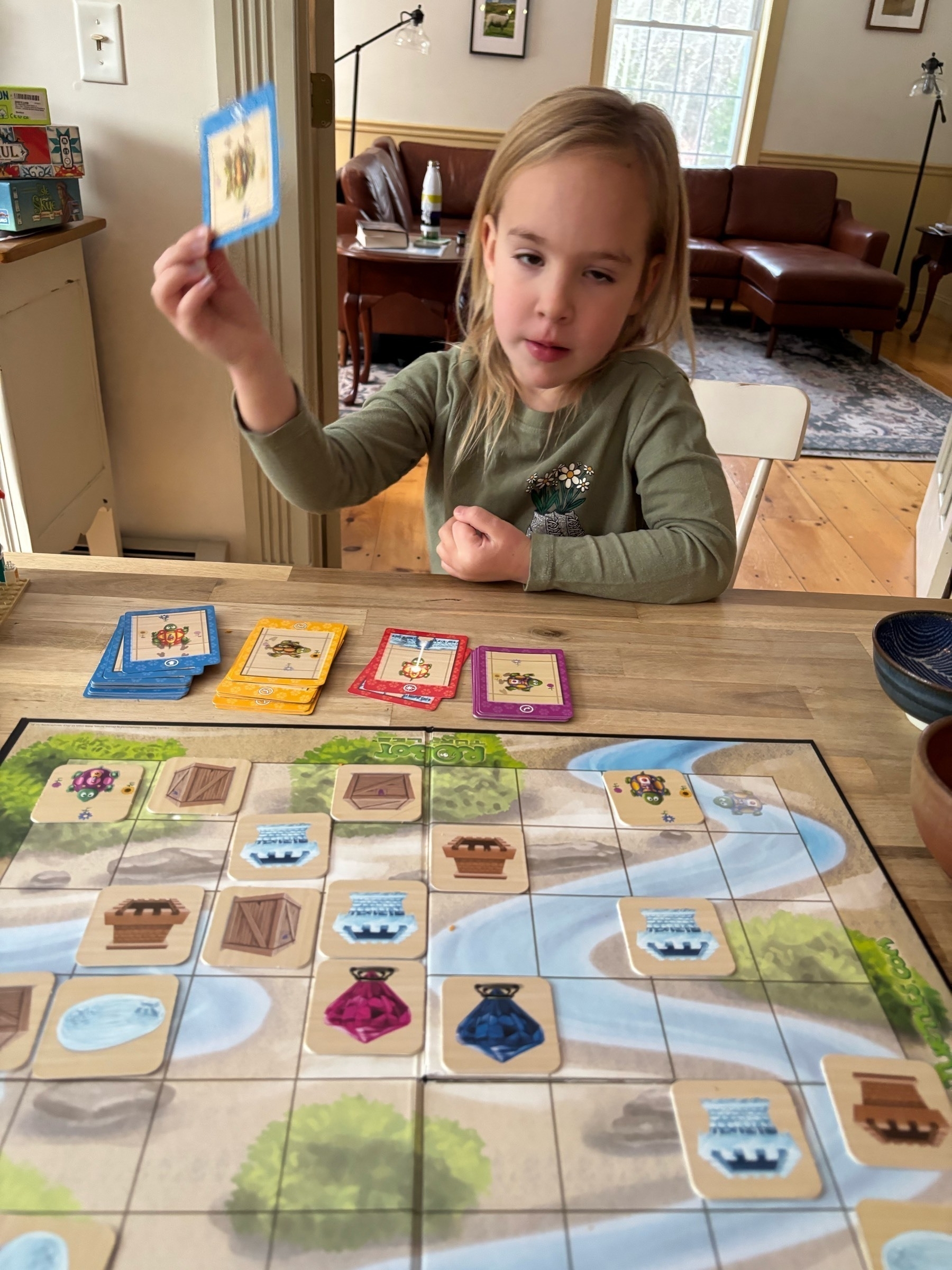 A young blond kid sits in front of the board game "robot turtles".  Blocks representing obstacles are in front of her, and she is handing over a card to help her turtle navigate the board.