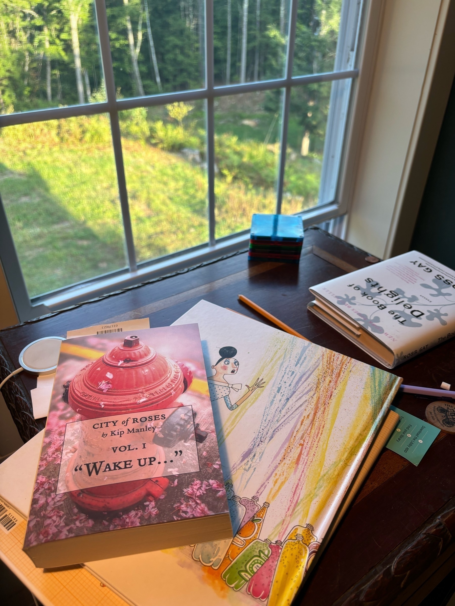 A book, with a fire hydrant on the cover, titled "City of Roses vol 1 'Wake Up...'" by Kip Manley, sits on a wooden table, on top of a children's book, and near Ross Gay's "Book of Delights".  Behind this are windowpanes and bright green grasses and trees