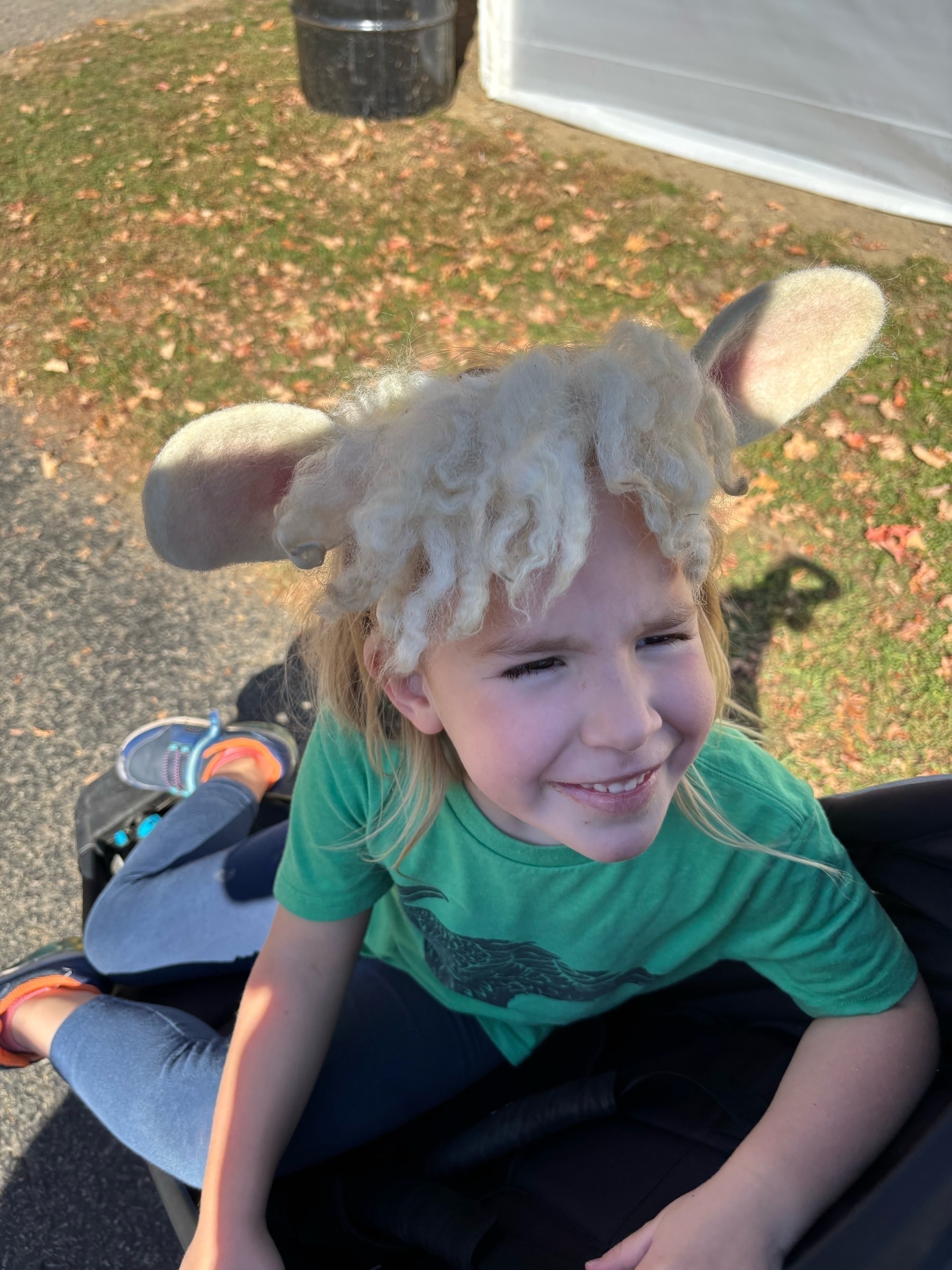 A white kid with blond hair grins up from a stroller. She's wearing a green shirt and sheep ears