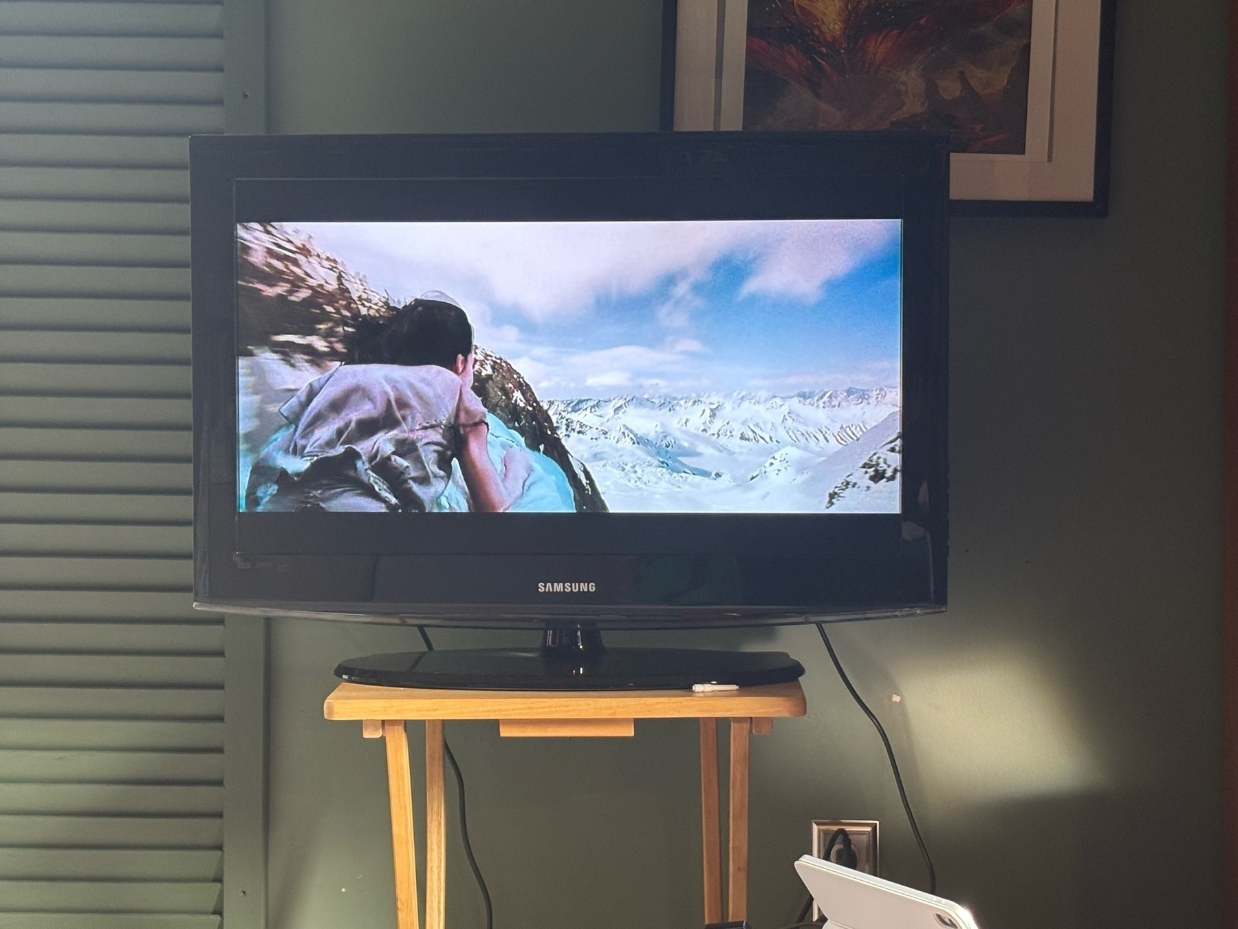 A picture of a television on a wooden table in front of a dark green wall. The TV is playing "Neverending Story" - looking from behind a boy with dark hair & a brown shirt sitting on the white dog-head of a luck dragon, flying over clouds