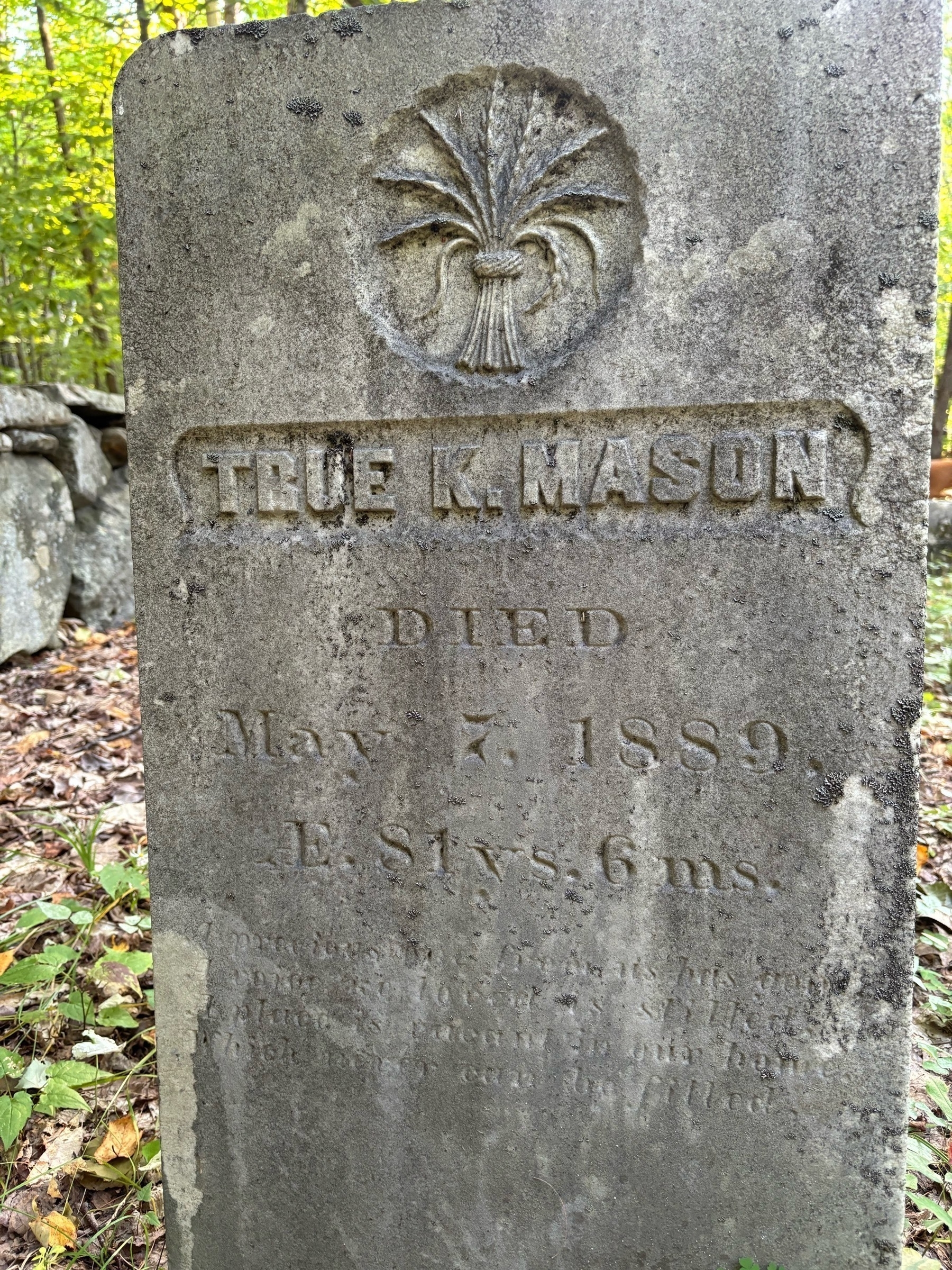 Close up of a weathered grey headstone.  It is a tall and thin rectangle with a carved sheaf of wheat on top, the name "True K Mason" below and date of death may 7, 1889.  Additional dates and a passage engraved on the stone below are not legible.