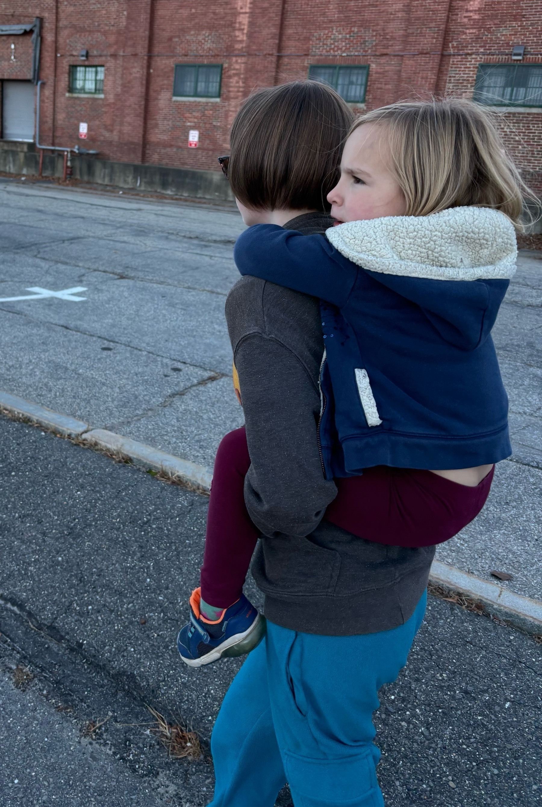 A teen in bright blue pants and freshly-cropped hair walks away from the camera with a young blonde kid in red pants and a puffy blue fleece carried piggyback.  A parking lot & brick building are in the background.