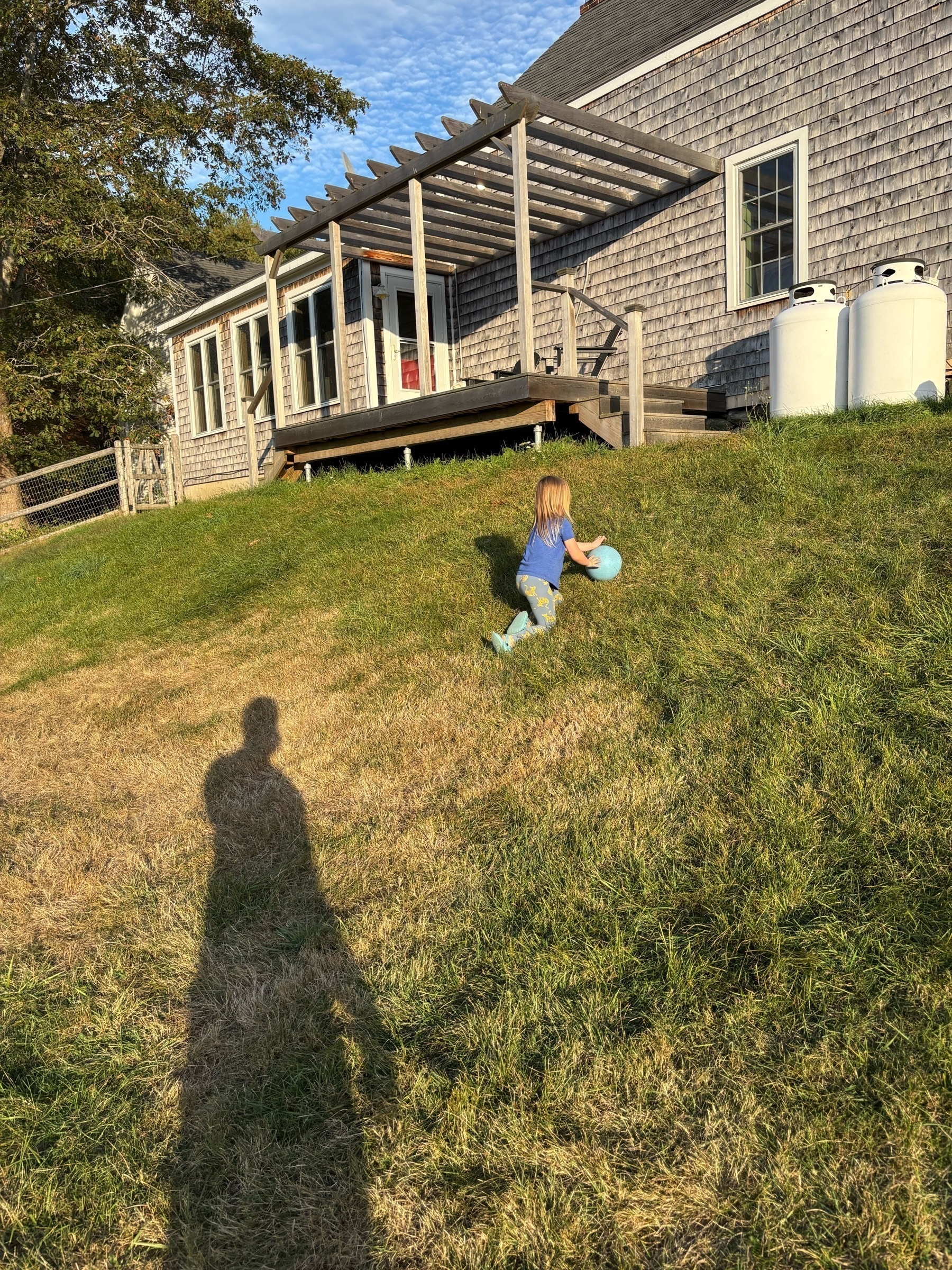 A delighted 6 year old with blond hair is on her knees rolling an awkward blue ball up a steep grassy hill with a wooden porch in the background.