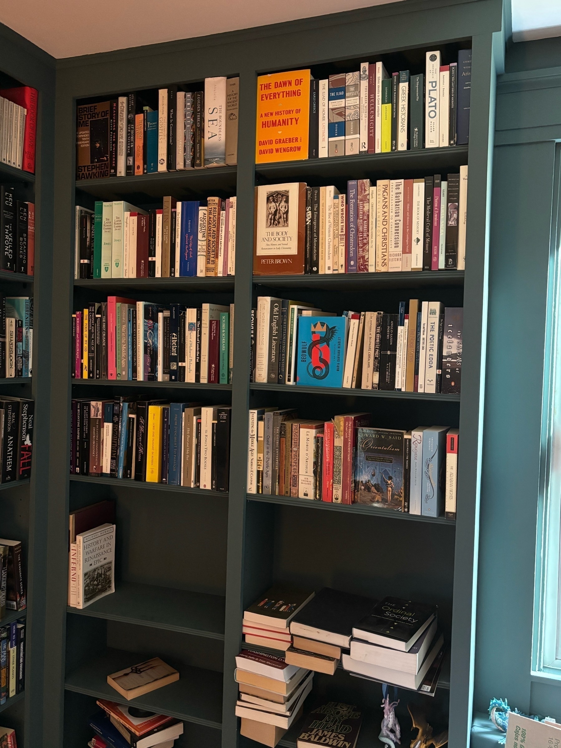 A set of bookshelves, some organized, others with piles and figurines on them.  Some books facing out include Peter Brown's Body and Society, Graeber and Wengrow's Dawn of Everything and Hawking's Brief History of Time