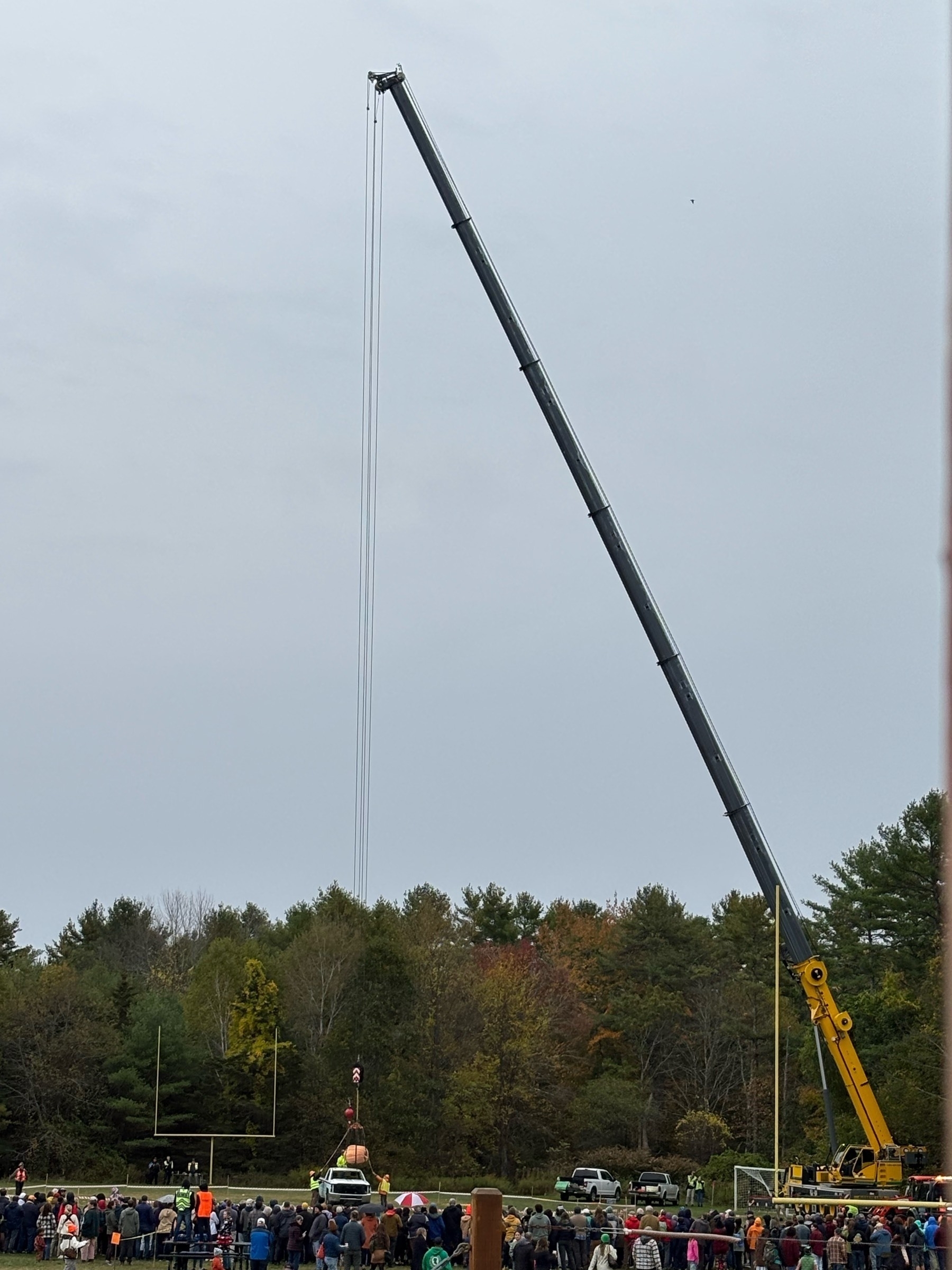 A crane holding a giant pumpkin just over a junked pickup truck in preparation for a huge pumpkin drop on a cloudy day.