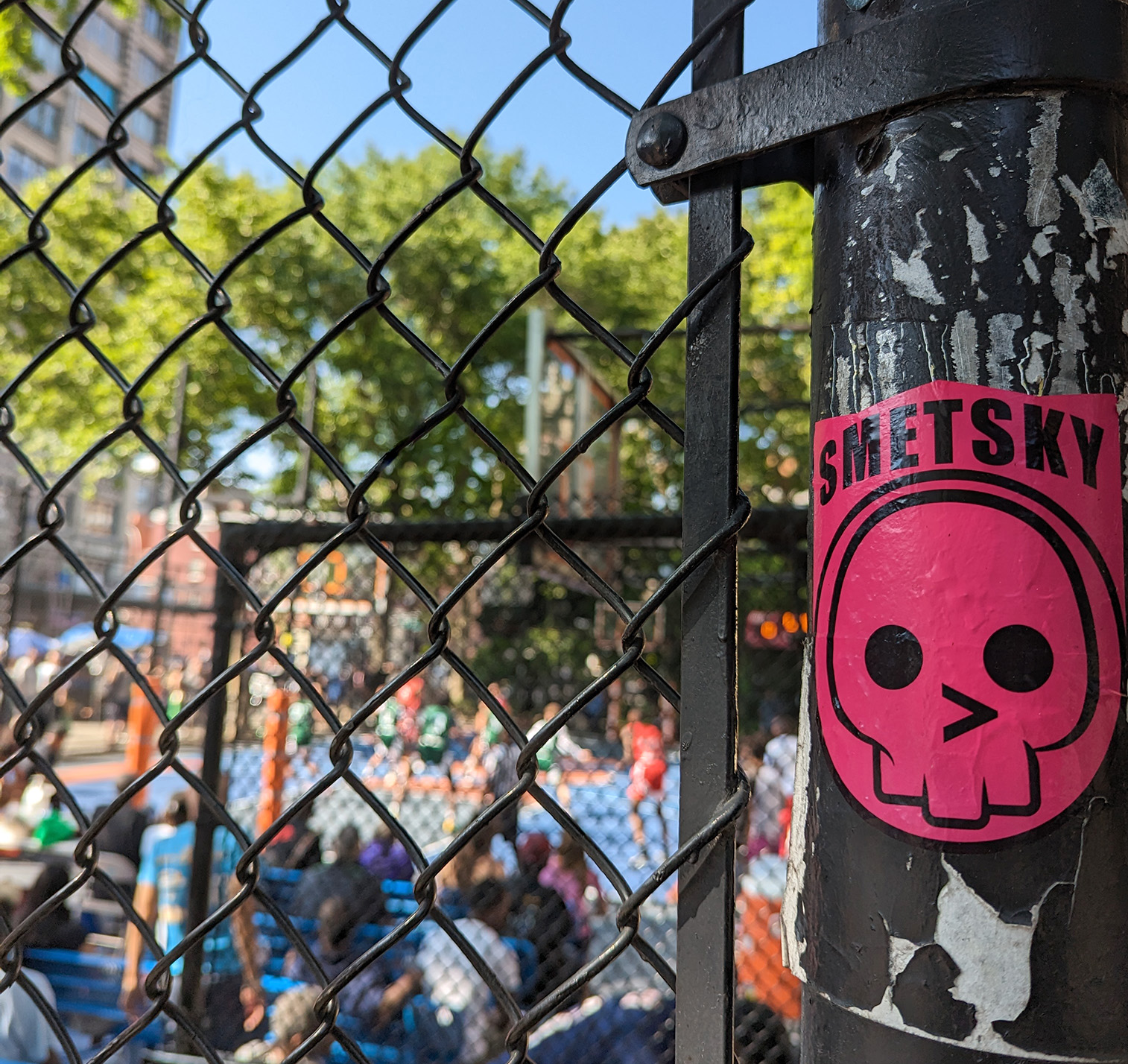 A black link fence in the foreground, with a pink sticker of a skull symbol. The sticker says Smetsky. In the background, a group of players in red and green uniforms playing basketball. An audience is watching.

