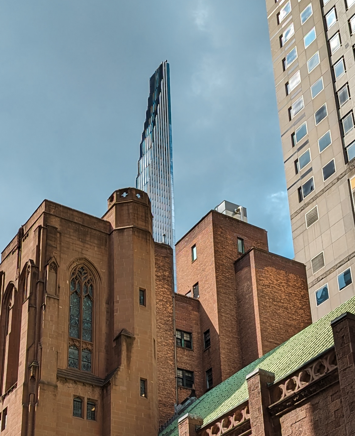 Steinway Tower, a narrow glass supertall skyscraper in NYC. In the foreground, a wall of a cathedral