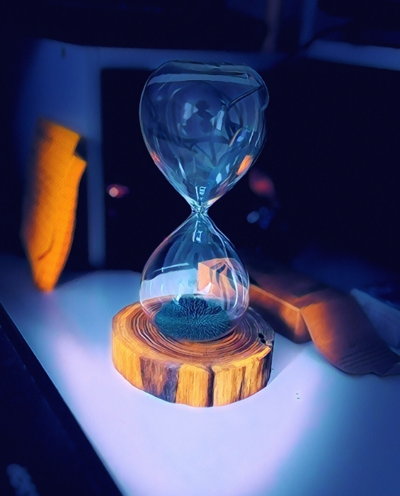An hourglass standing on a desk on a wooden plastform. In the background some electronics and wires