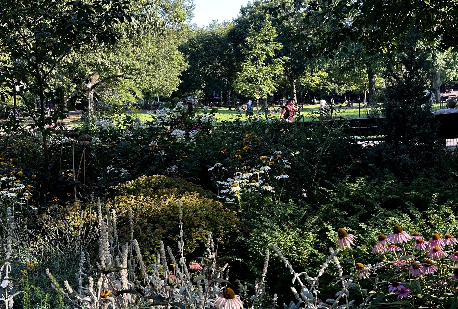 A picture of a park. People walking up and down a path, flowers in the foreground and different colors, green grass in the background with some trees. &10;