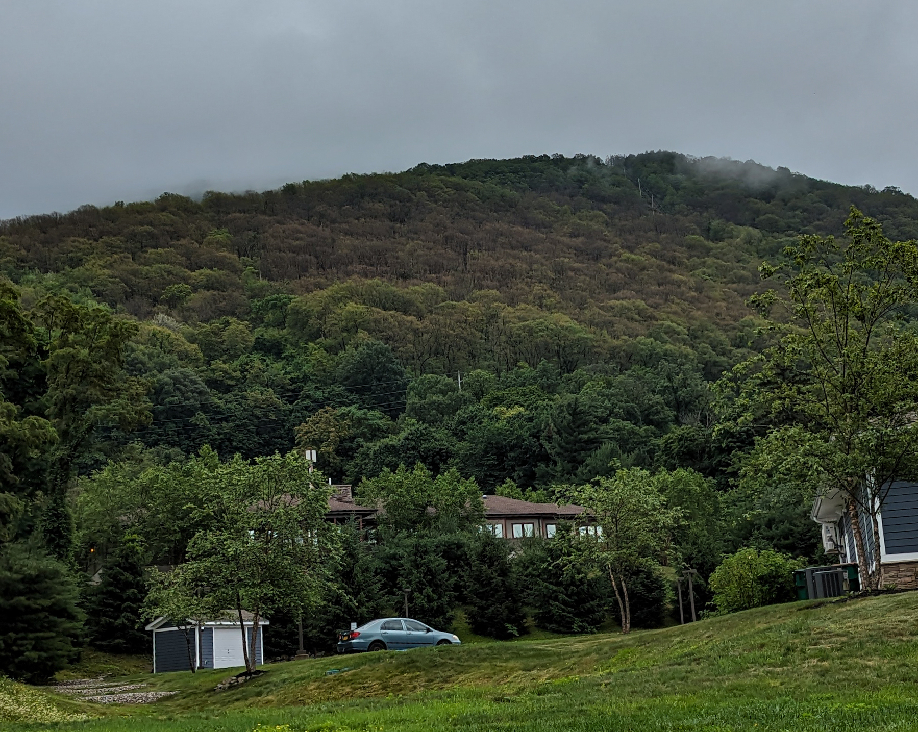 Mount Beacon, from the town level. The mountain is covered with trees all the way to the top, where It's covered with a bit of a low grey cloud. There's a car and a house in the foreground. 
