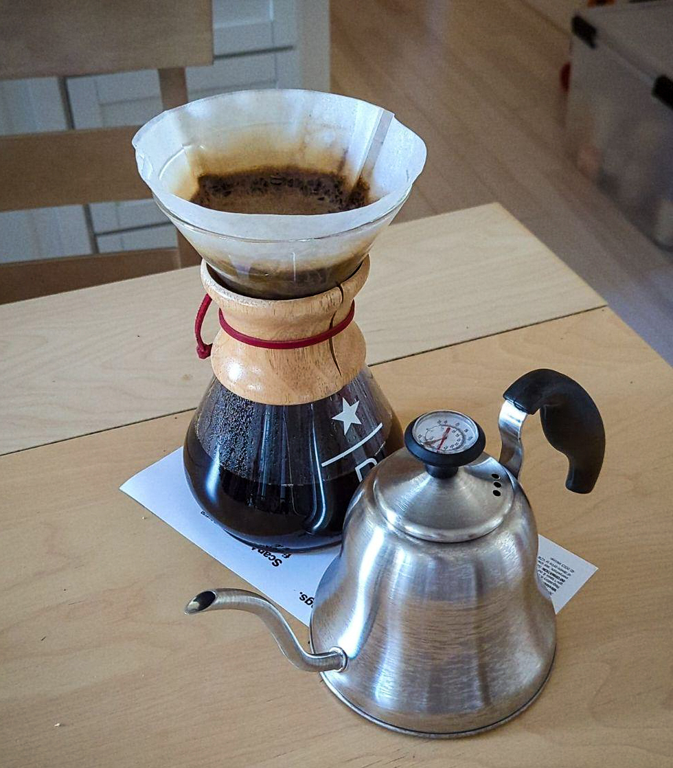 A chemex pour over loaded with coffee and hot water in a filter on top, next to a small hotpot with a long snot and a thermometer. They are both on a wooden kitchen table. 