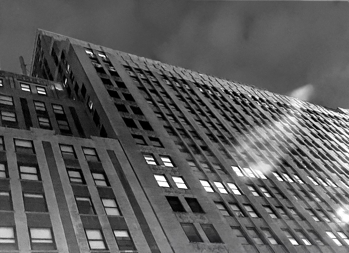 A black and white photo of a New York City office building at night, with some windows lit and most dark. A fluff of smoke is caught in the foreground.