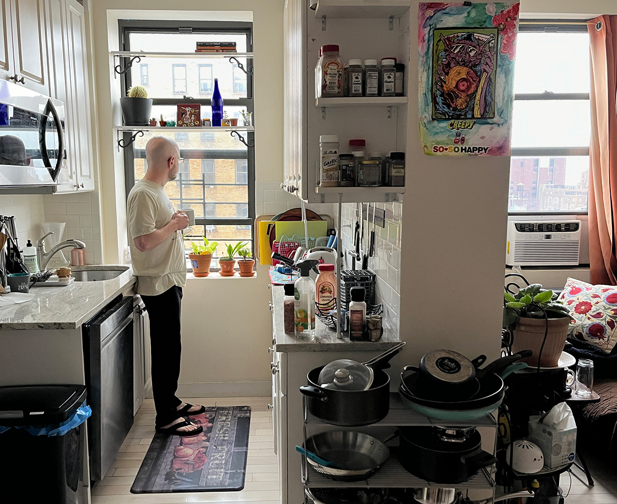 A picture of a small manhattan apartment. Standing at the left, a bald guy with a cup in his hand looking outside. The apartment is full of objects, spices, a sofa, a table, a microwave, and two windows looking outside at the gloomy grey day.
