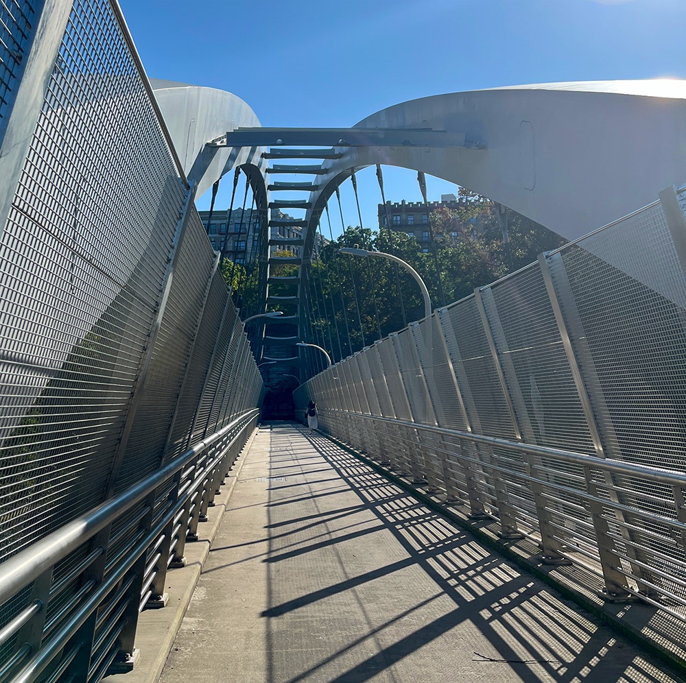 Crossing a metallic bridge, with the sun at the upper left. The railing are curved and stylish. A woman is walking ahead.
