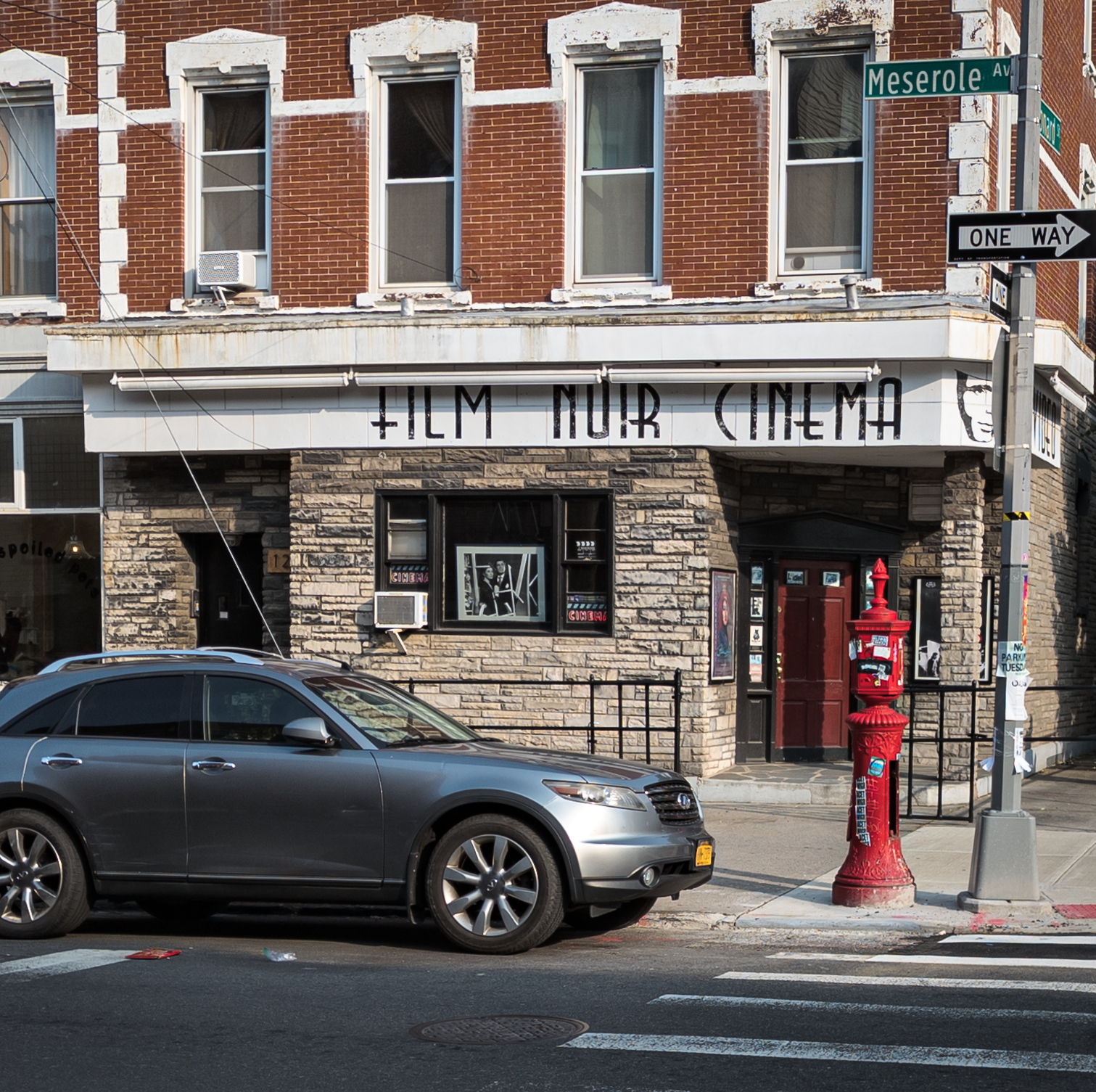 The store, Film Noise Cinema, on the street, with the name in large black letter hanging down. A red door leads inside, it's closed. There's a car parked in front and a red NYC emergency phone for firefighters in front of it