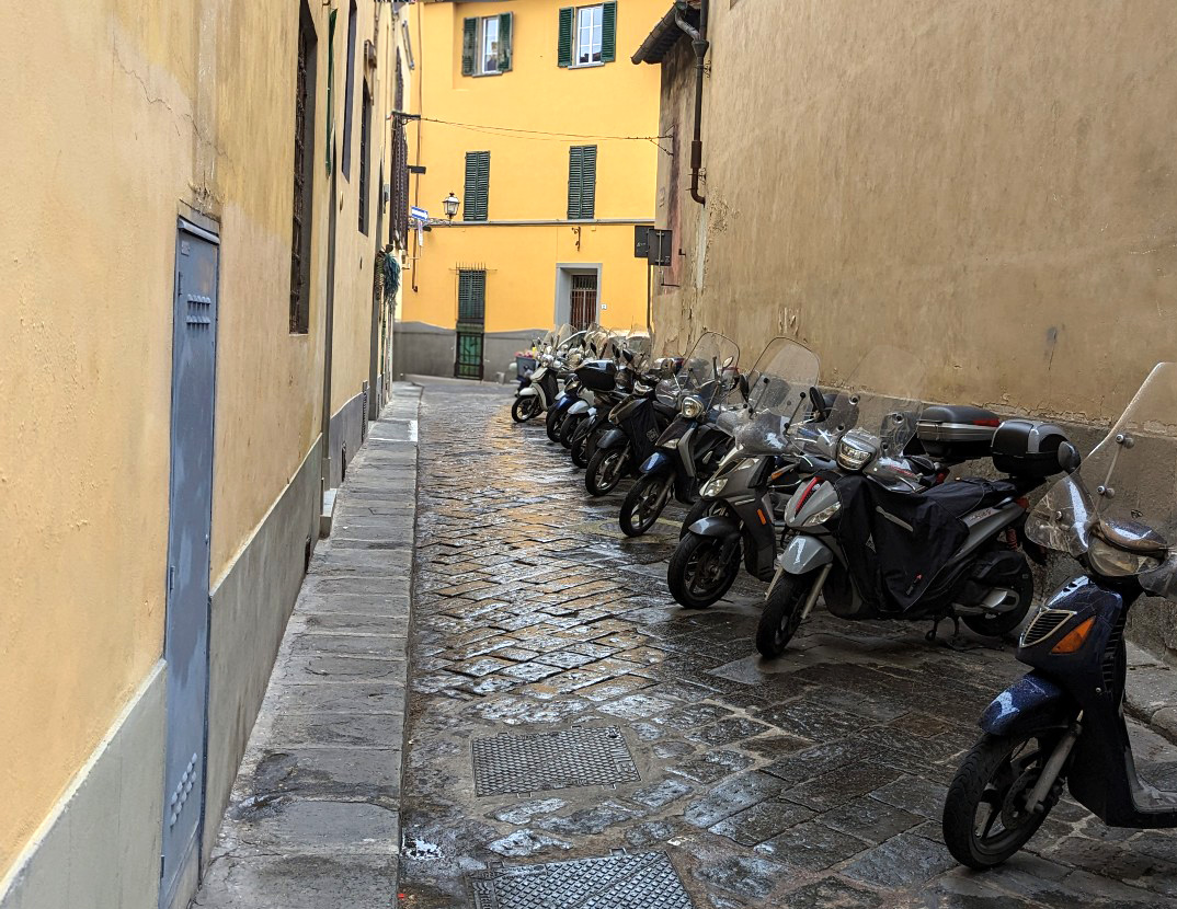 A paved road with a row of scooters next to each other blocking about half of the street. Yellow-painted walls are closing in on the street, making it more of an alleyway. 
