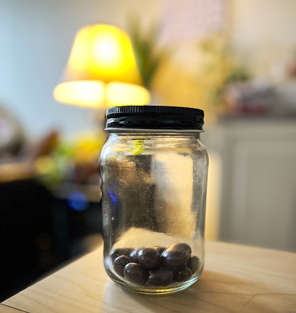 A glass jar filled with chocolates of different flavors, as expressed above. There’s a lamp in the background next to a Snake plant, providing yellow-orange light