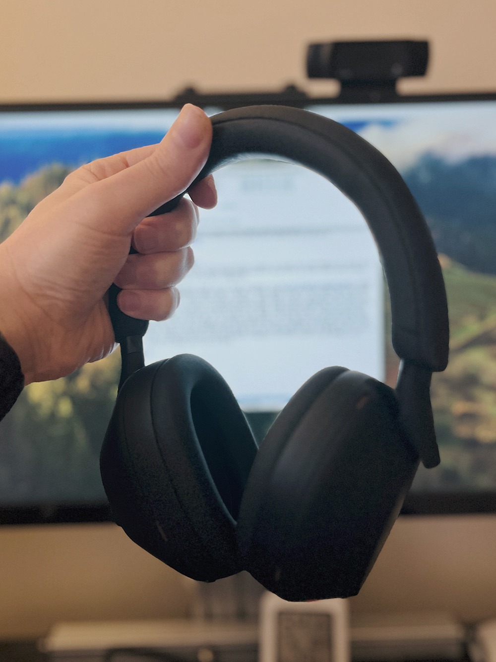 The Sony WH-1000XM5. An all-black, over the ear headset. My hand holds it by the head band, my screen blurred in the background. 