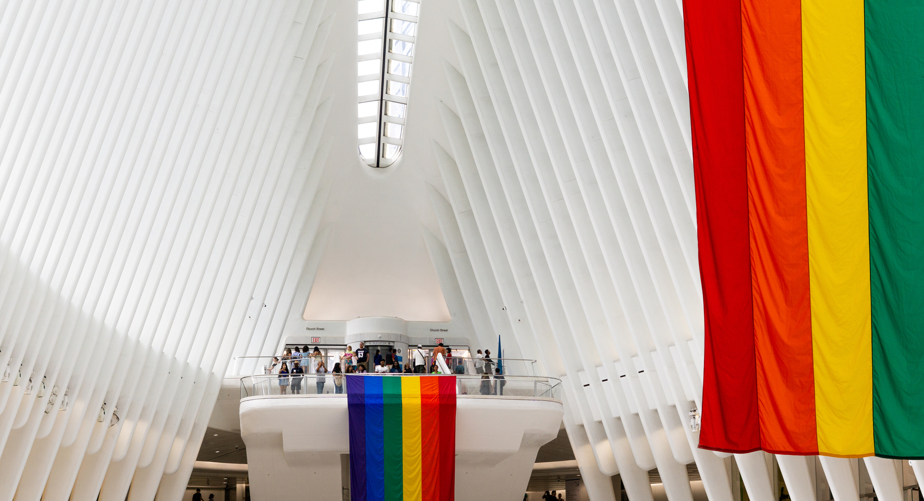 The inside of the Wesfield World Trade Center, where the white beams that look like the ribs of a giant dinosaur can be seen. There are two pride flags; one at the foreground at the right side of the photo, and one at the center, hanging from one of the balconies inside