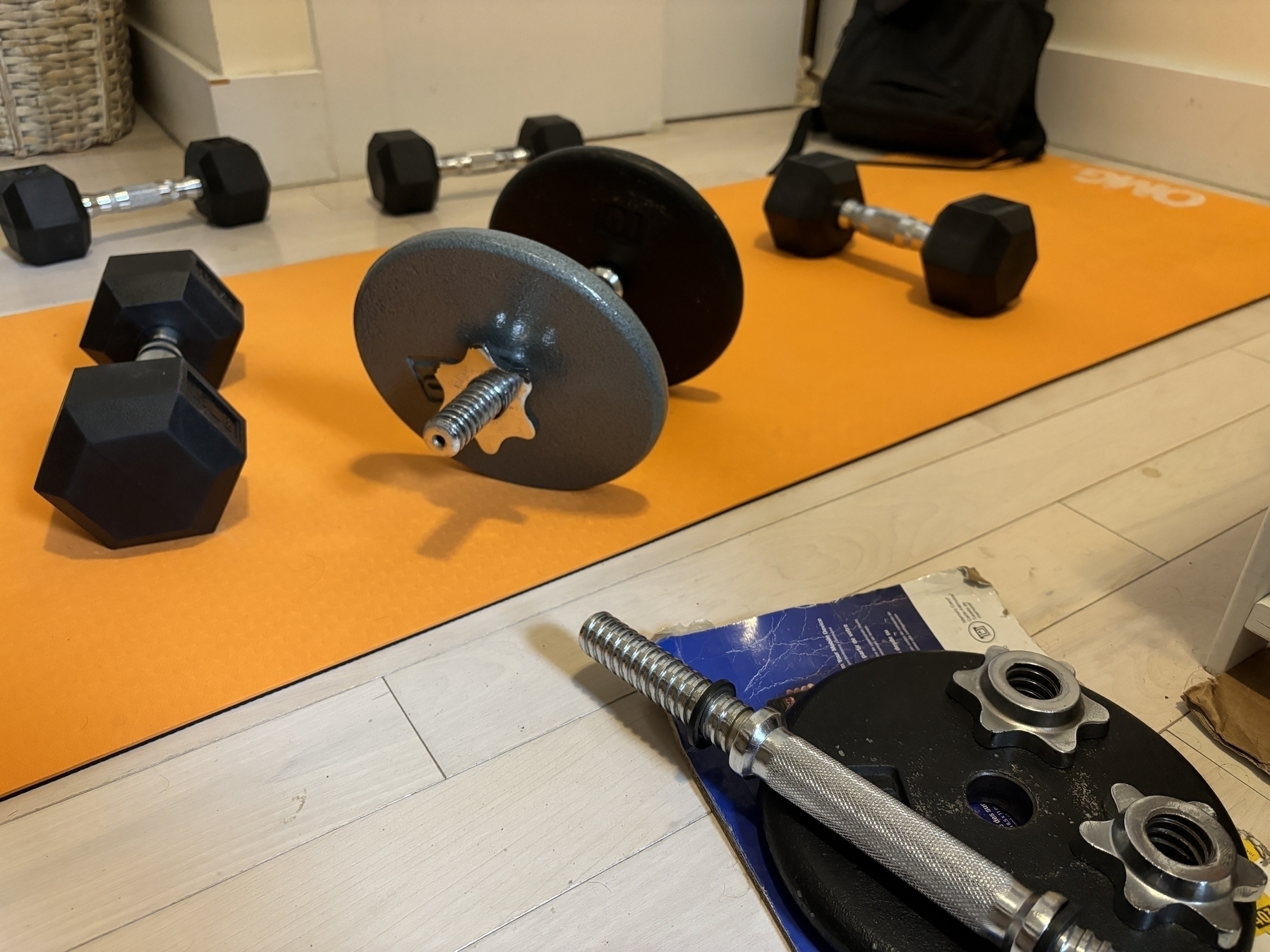 Auto-generated description: A home workout setup with various dumbbells and weight plates on an orange exercise mat.