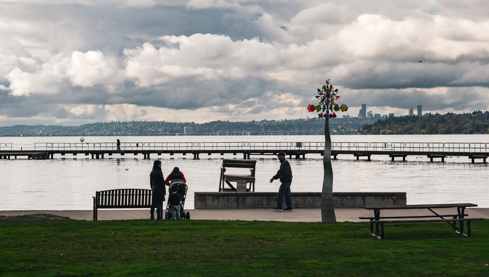  A waterfront park with people standing by benches with a cloudy sky overhead and distant views of a pier and city skyline. There's a vibrant colorful artificial statute next to the people.