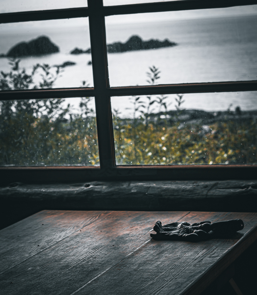 Auto-generated description: A wooden table is set near a window looking out at islands on a misty sea, with a pair of gloves resting on the table.