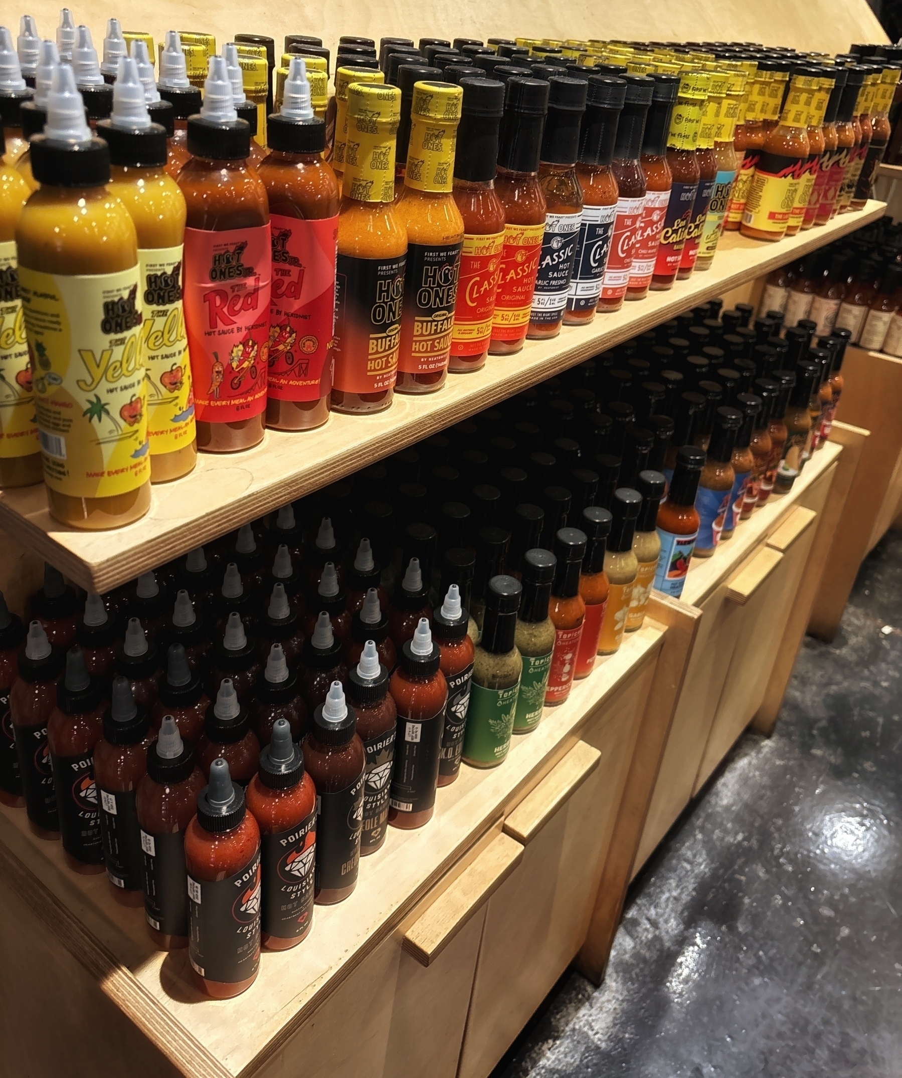 Two rows of hot sauces in various colors on wooden shelves