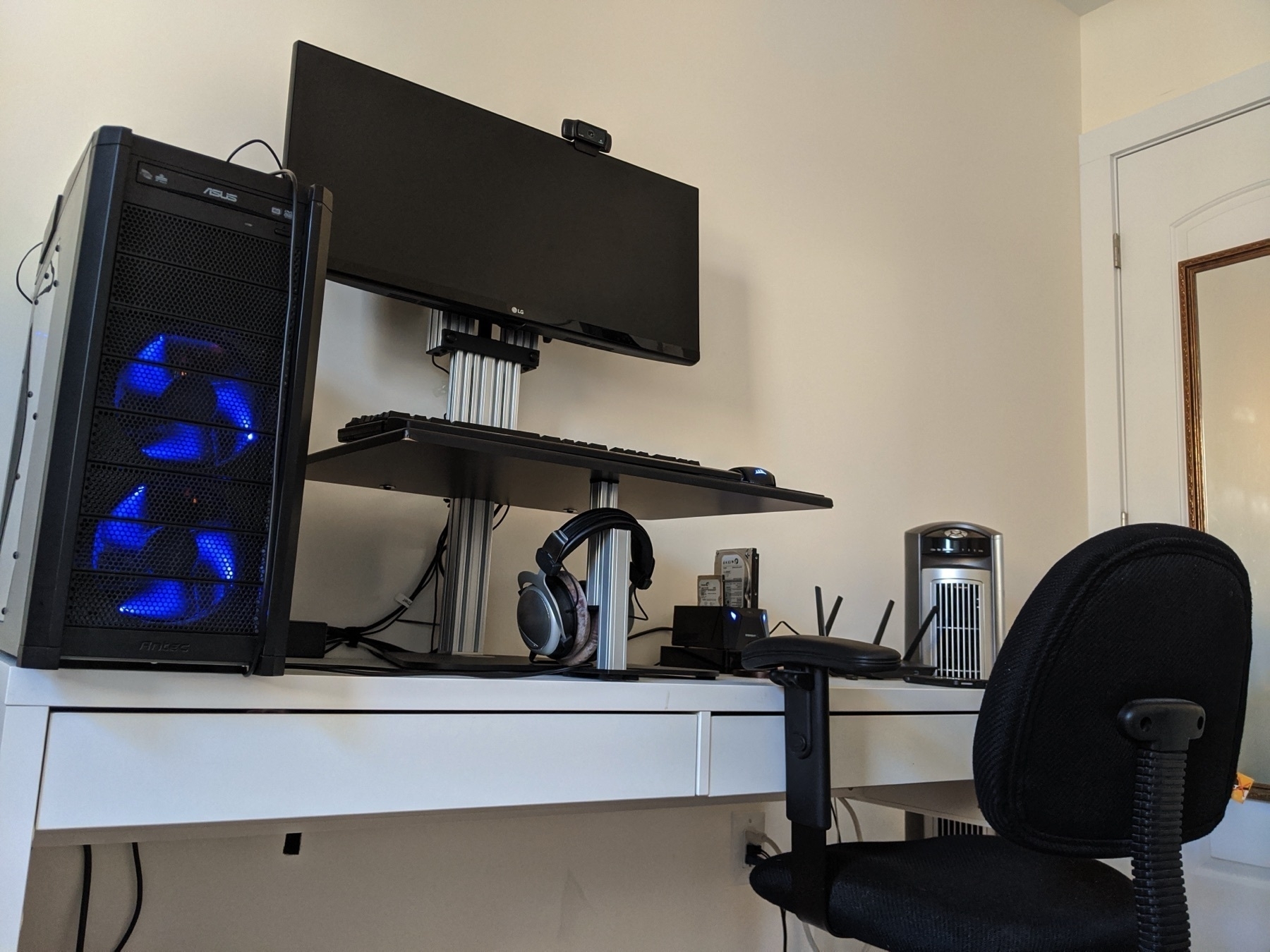 A home office setup features a monitor stand, desktop computer with blue-lit fans, headphones, a router, and a webcam, all on a white desk with a black office chair.